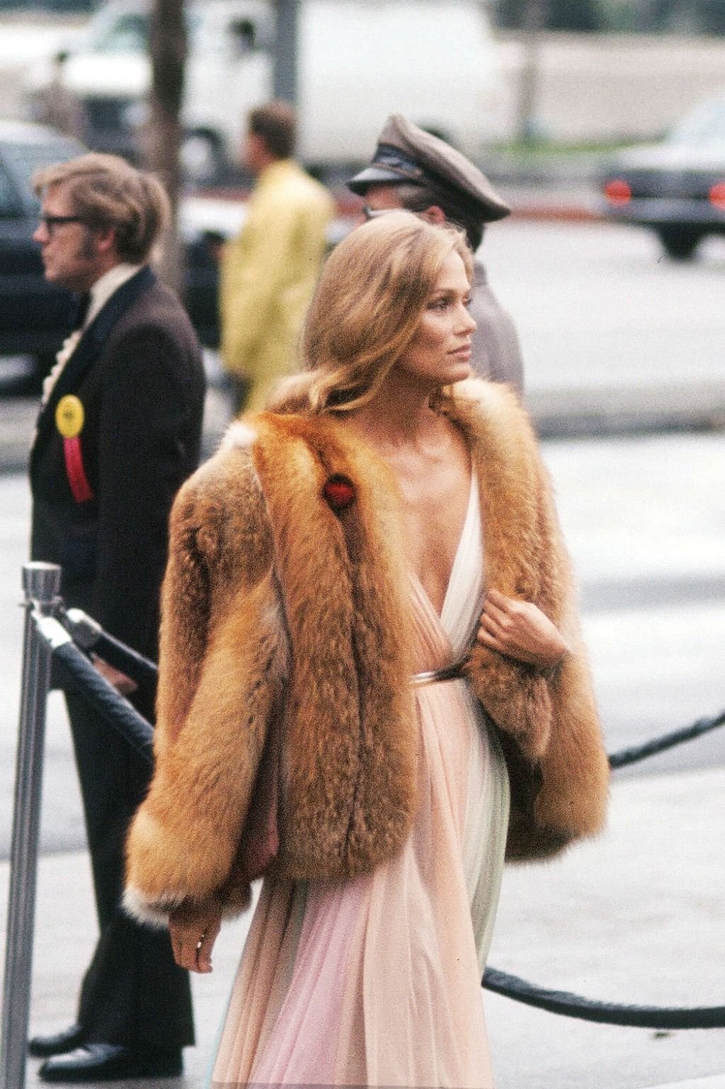 Lauren Hutton in a light-colored gown and a large fur coat walking outdoors, with people and cars in the background.