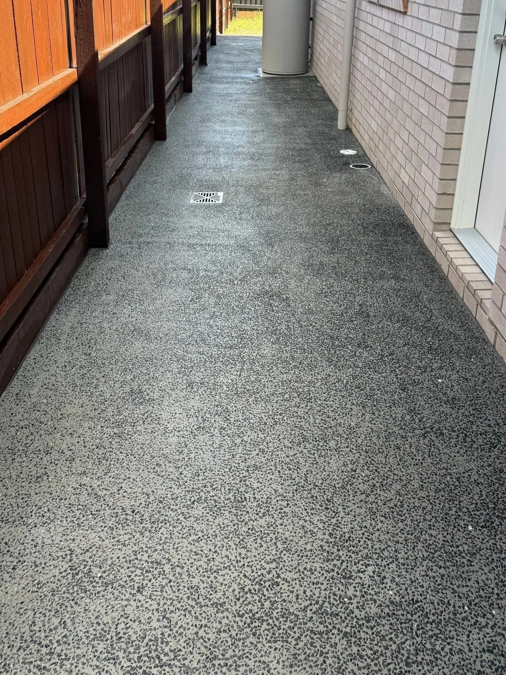 A narrow outdoor patio with a speckled concrete floor, brick and wood fencing, and various drainage covers.