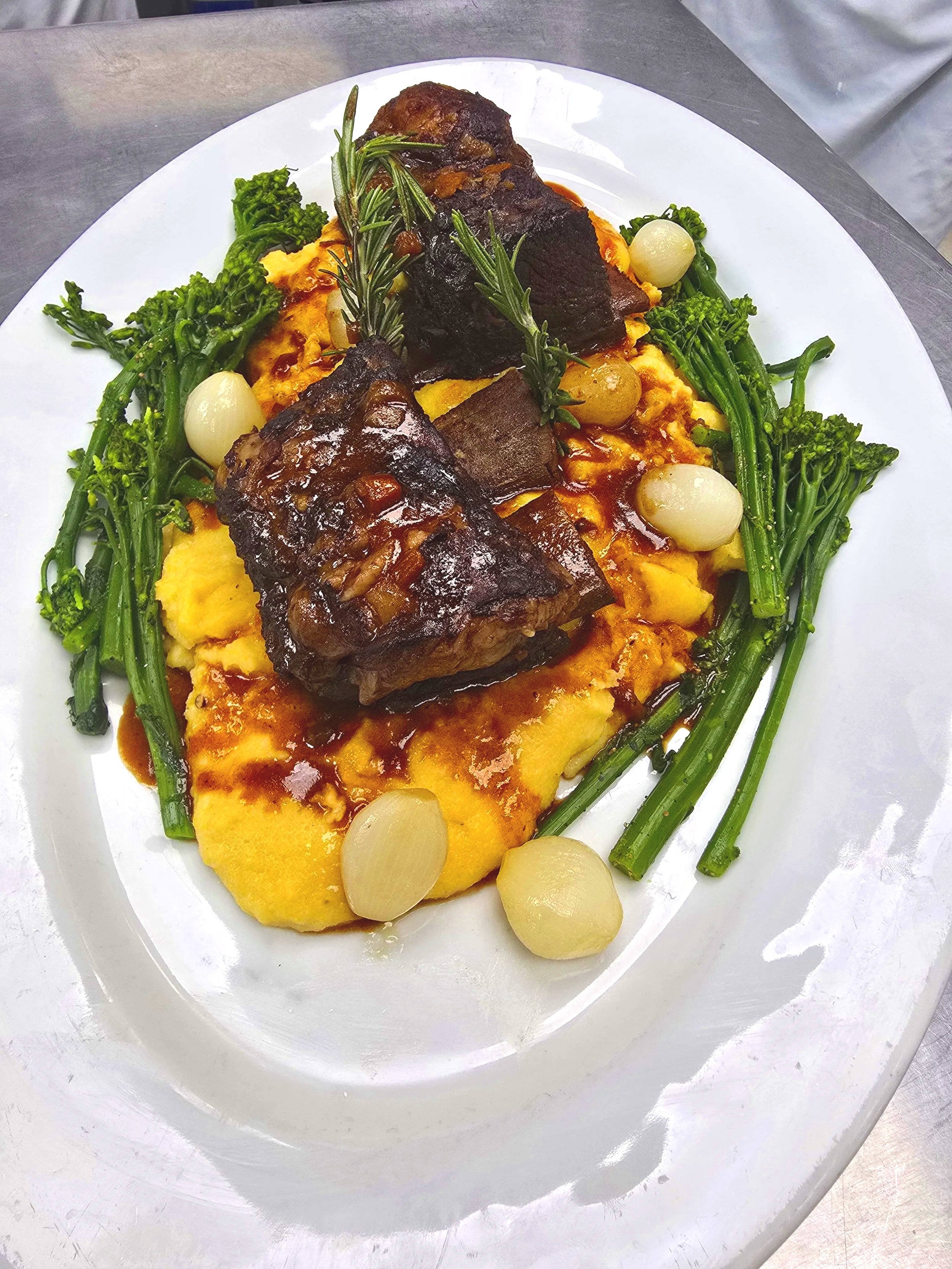 Plate of food with beef ribs, scrambled eggs, broccolini, pearl onions, and sprigs of rosemary garnished on a white dish.