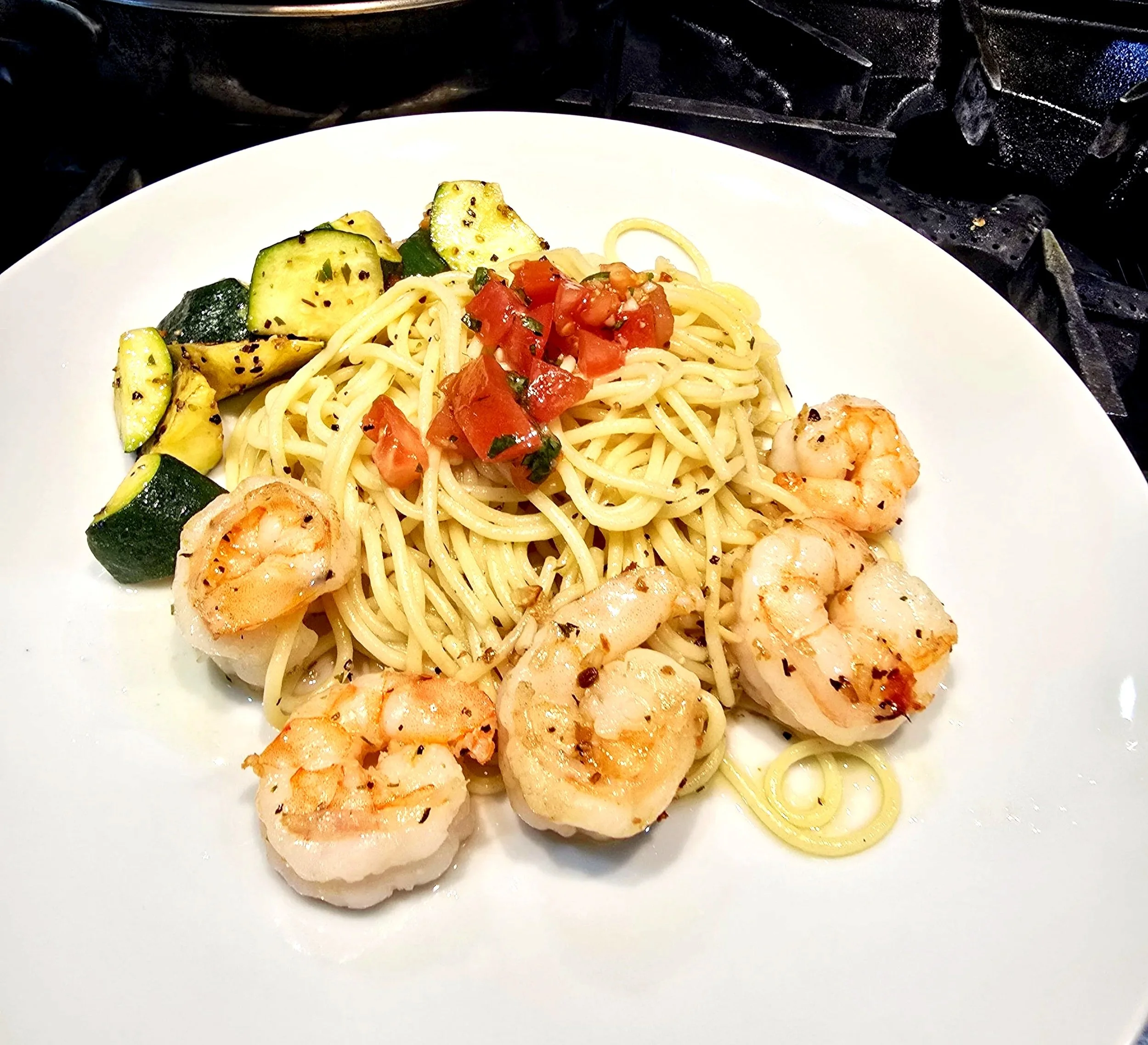 A white plate with cooked spaghetti topped with cherry tomatoes, grilled zucchini, and shrimp garnished with herbs, placed on a stove.