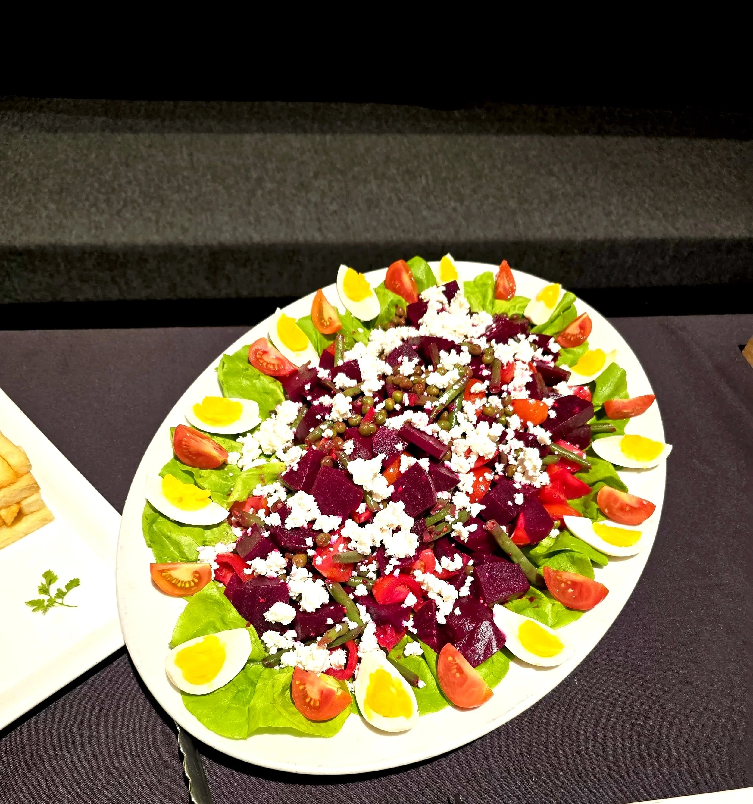 Colorful plated salad with cherry tomatoes, hard-boiled egg slices, beets, green beans, crumbled cheese, and lettuce, served on a white dish against a dark background.
