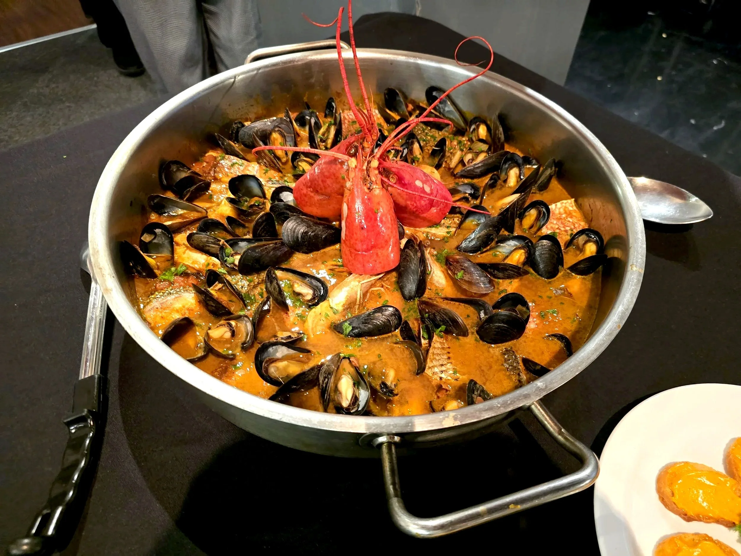 A seafood dish in a silver pan featuring a cooked lobster with mussels and a spicy broth, garnished with herbs. Part of a plate with sliced oranges is visible in the foreground.