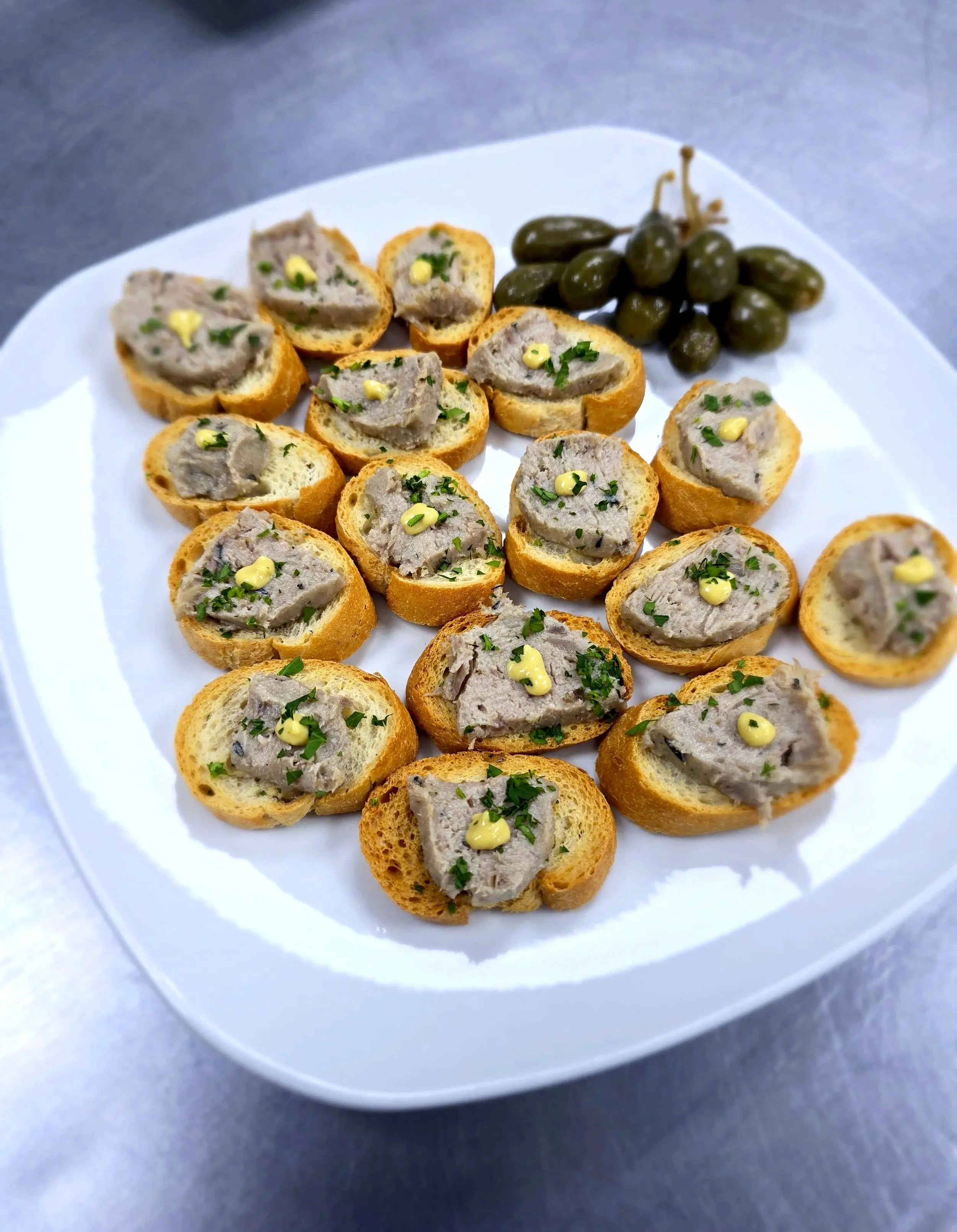 Plate of sliced baguette topped with pâté, garnished with chopped herbs and small dollops of mustard, served with a bunch of green grapes.