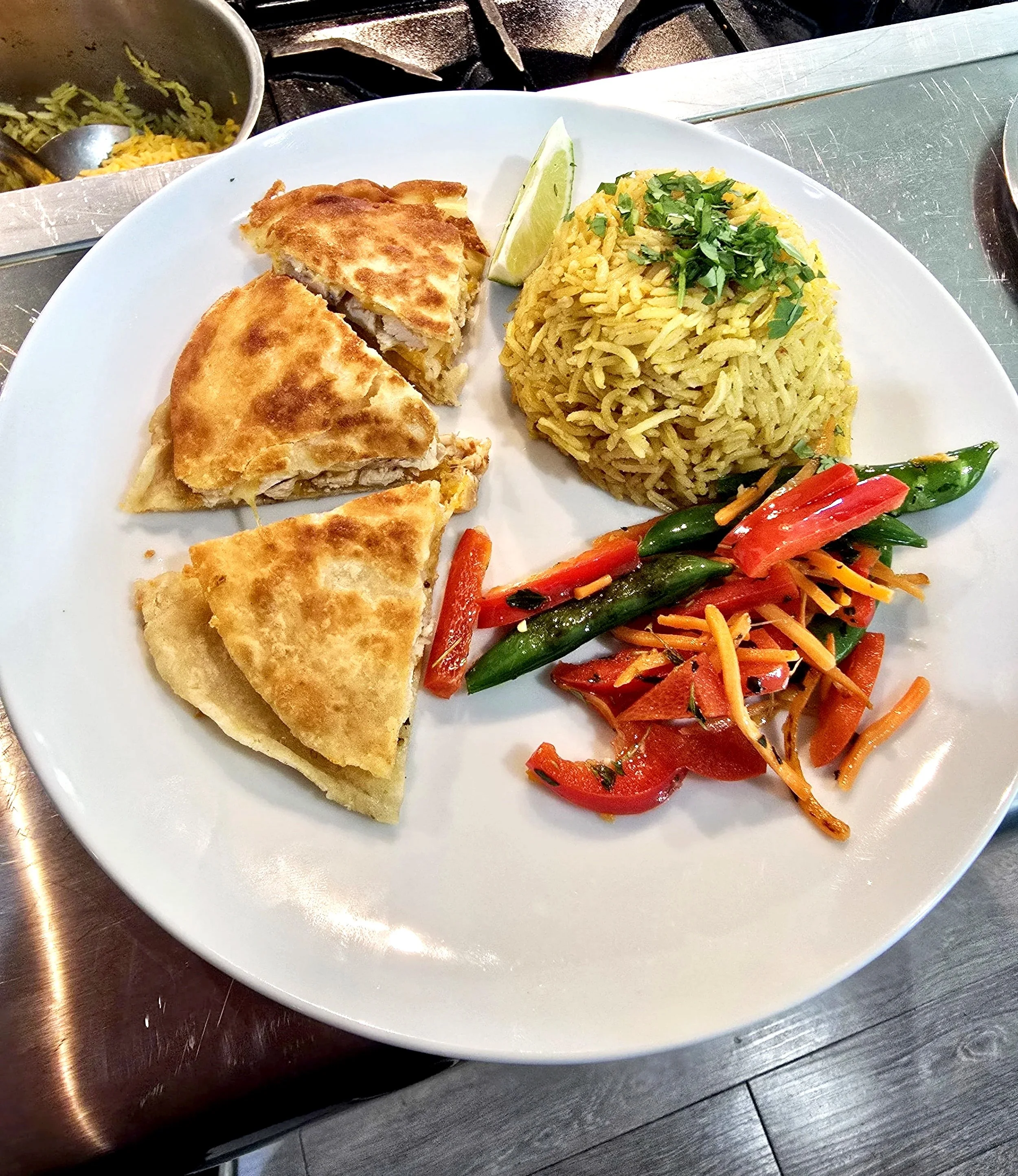 A white plate with rice, chicken pie, and mixed vegetable salad with carrots and peppers, garnished with chopped cilantro and lime wedge, on a stainless steel counter.