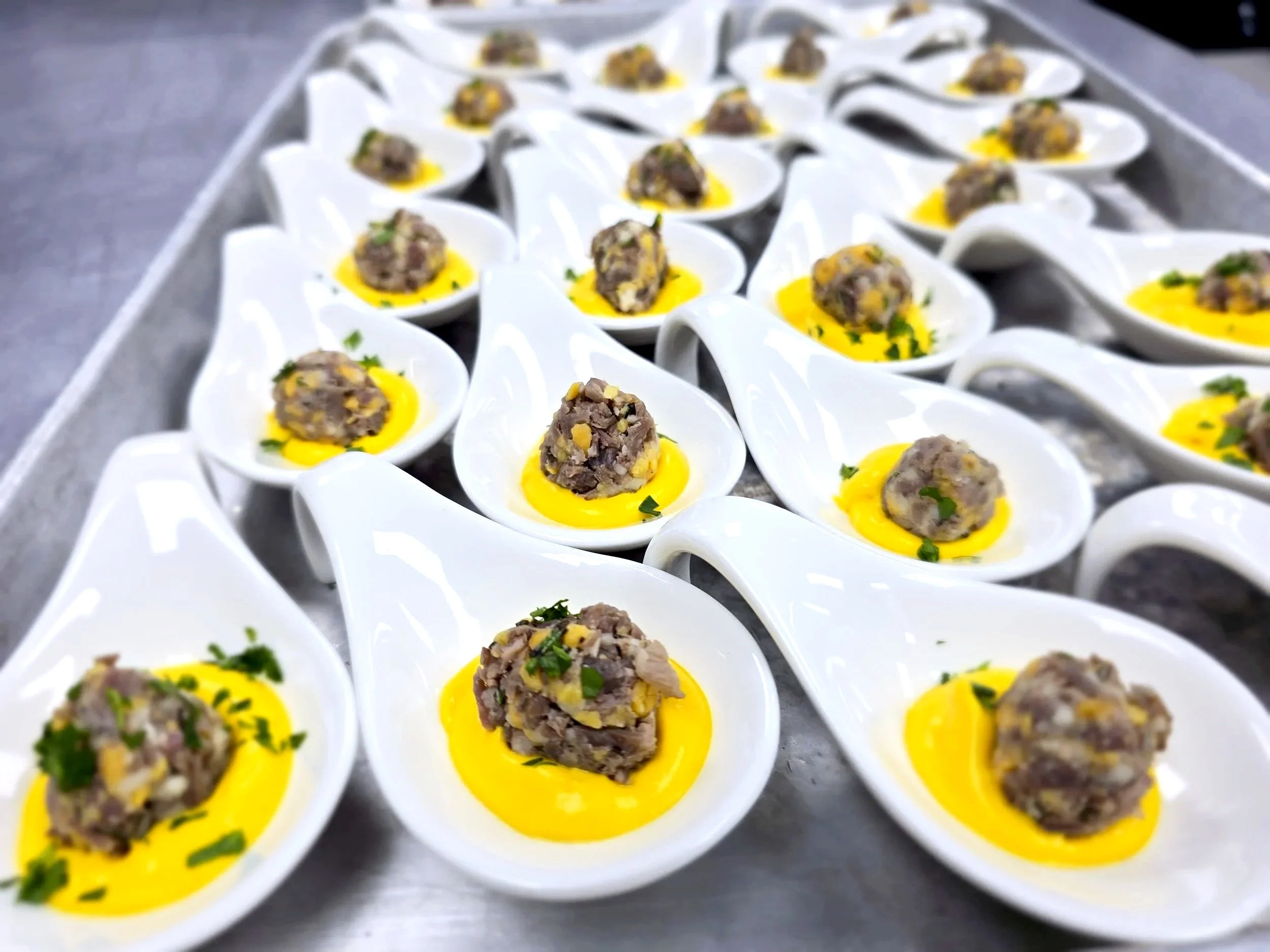 Small white spoons filled with a meat mixture and garnished with chopped herbs, placed on a tray with a yellow sauce underneath.