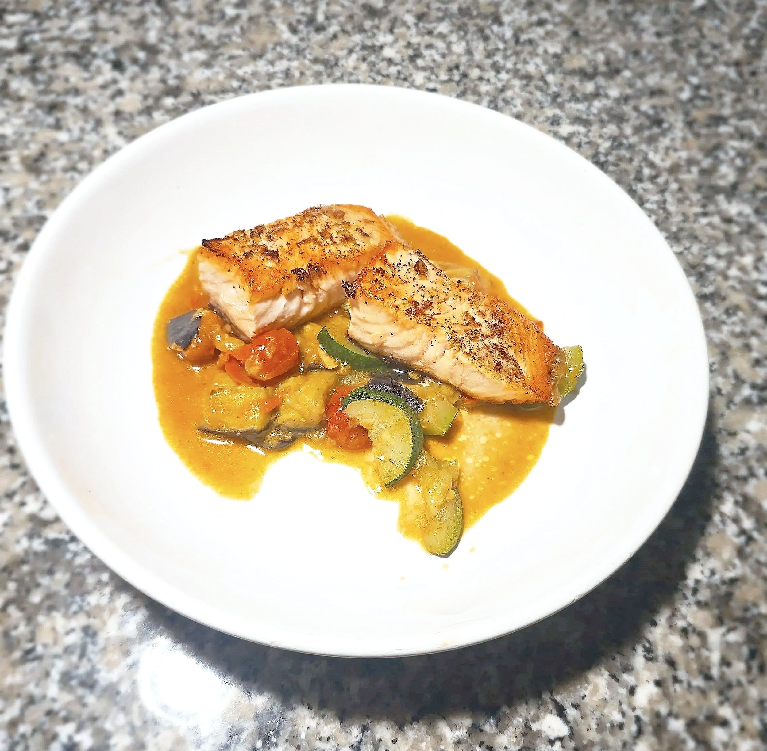 A white bowl containing cooked salmon fillet topped with black pepper, served with a yellow curry sauce with vegetables, on a speckled gray countertop.