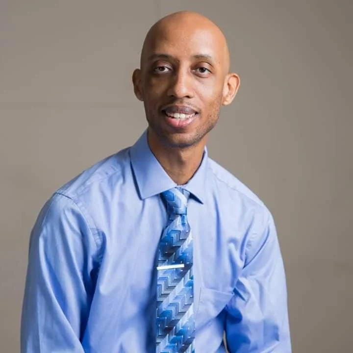 Headshot of a man with a bald head, light beard, wearing a light blue dress shirt and a patterned blue tie, smiling at the camera against a plain background.