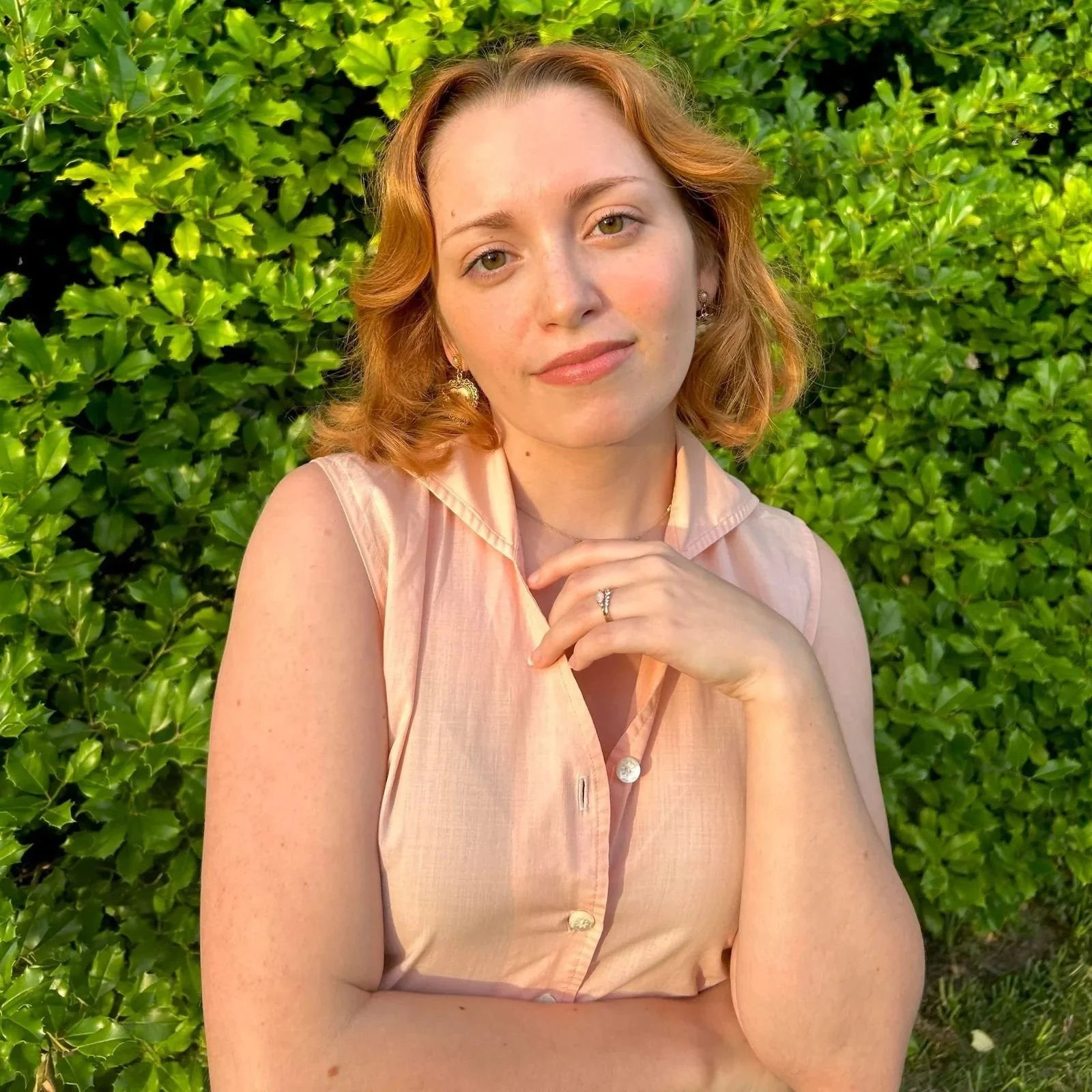 A woman with light skin, red hair, and green eyes posing in front of green leafy bushes, wearing a pink sleeveless blouse and earrings. Queer couples therapist in St. Louis