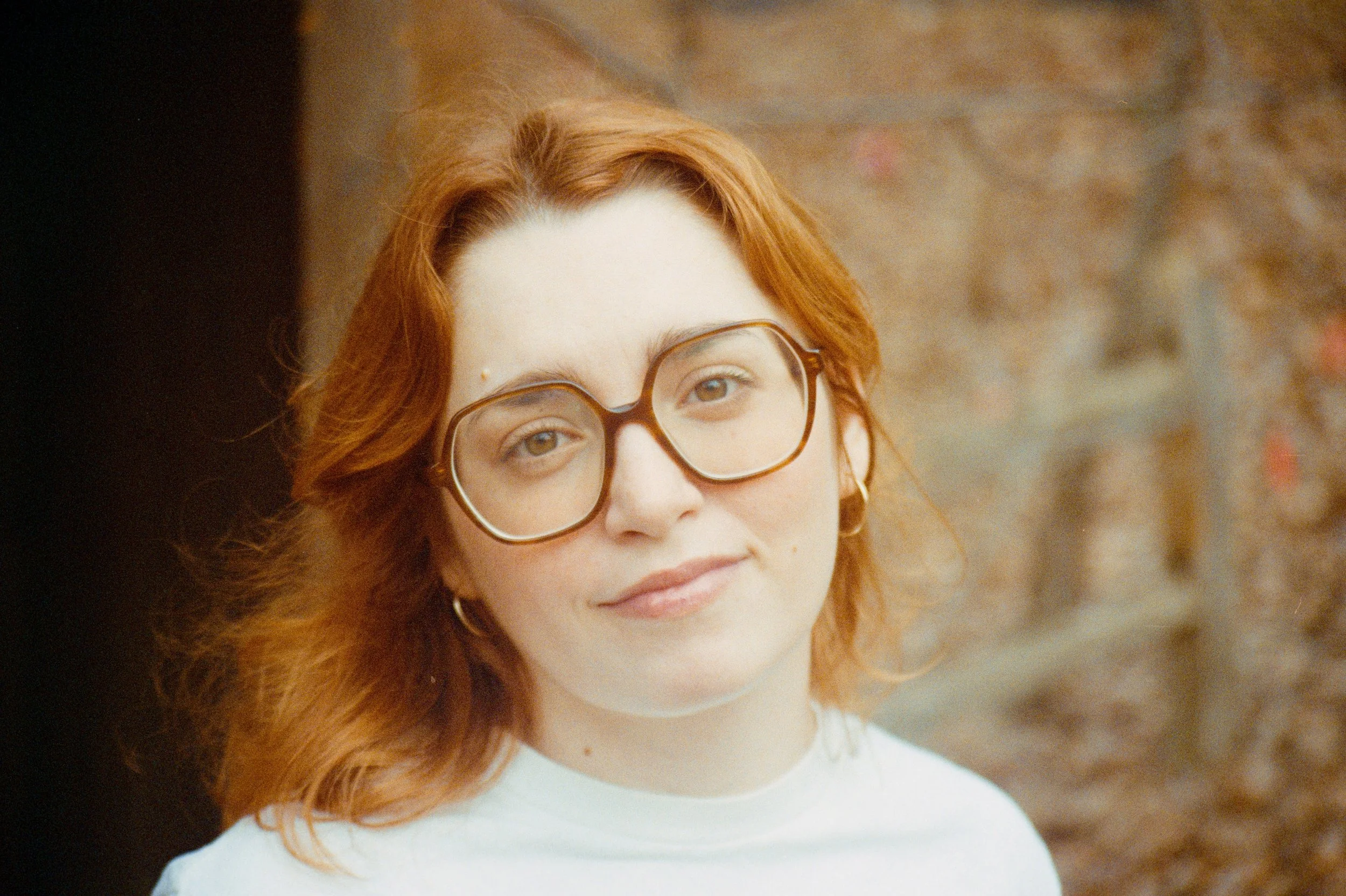 A woman with red hair and glasses looking at the camera, standing outdoors with a blurred background.