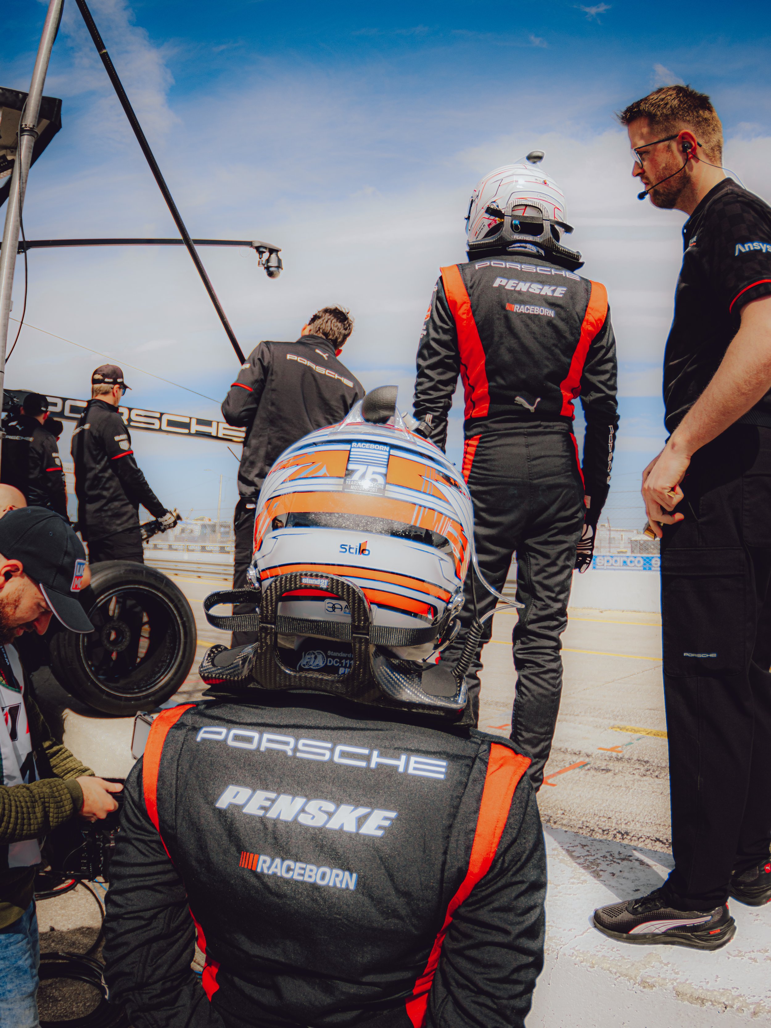 Race car driver team members in black and red uniforms preparing a race car in a pit lane, with the driver wearing a white helmet and only the back visible. The team members are working on the car and communicating.
