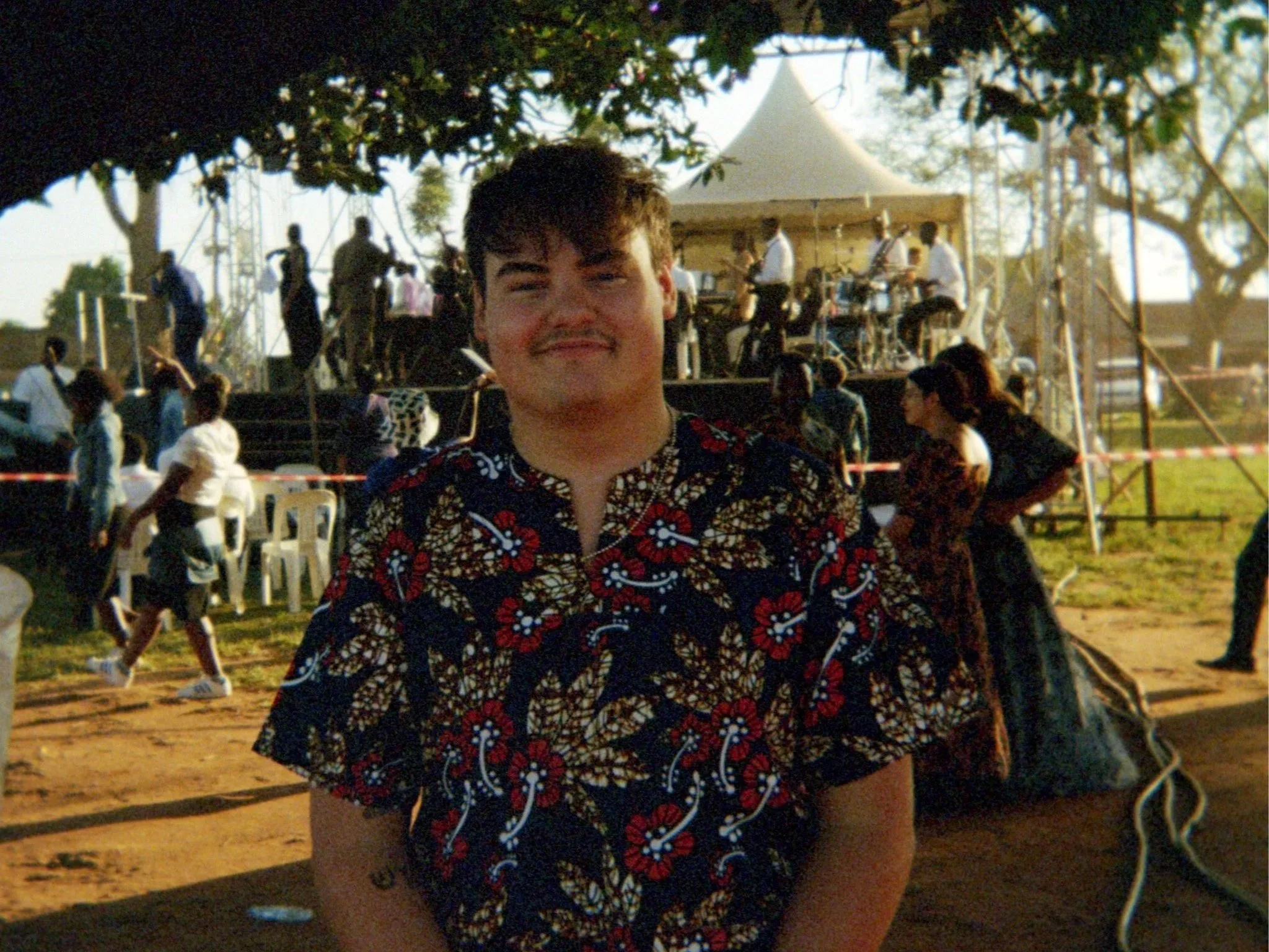 A young man smiling in front of a stage with a band playing, under a tent at an outdoor event, with other people sitting and standing around.
