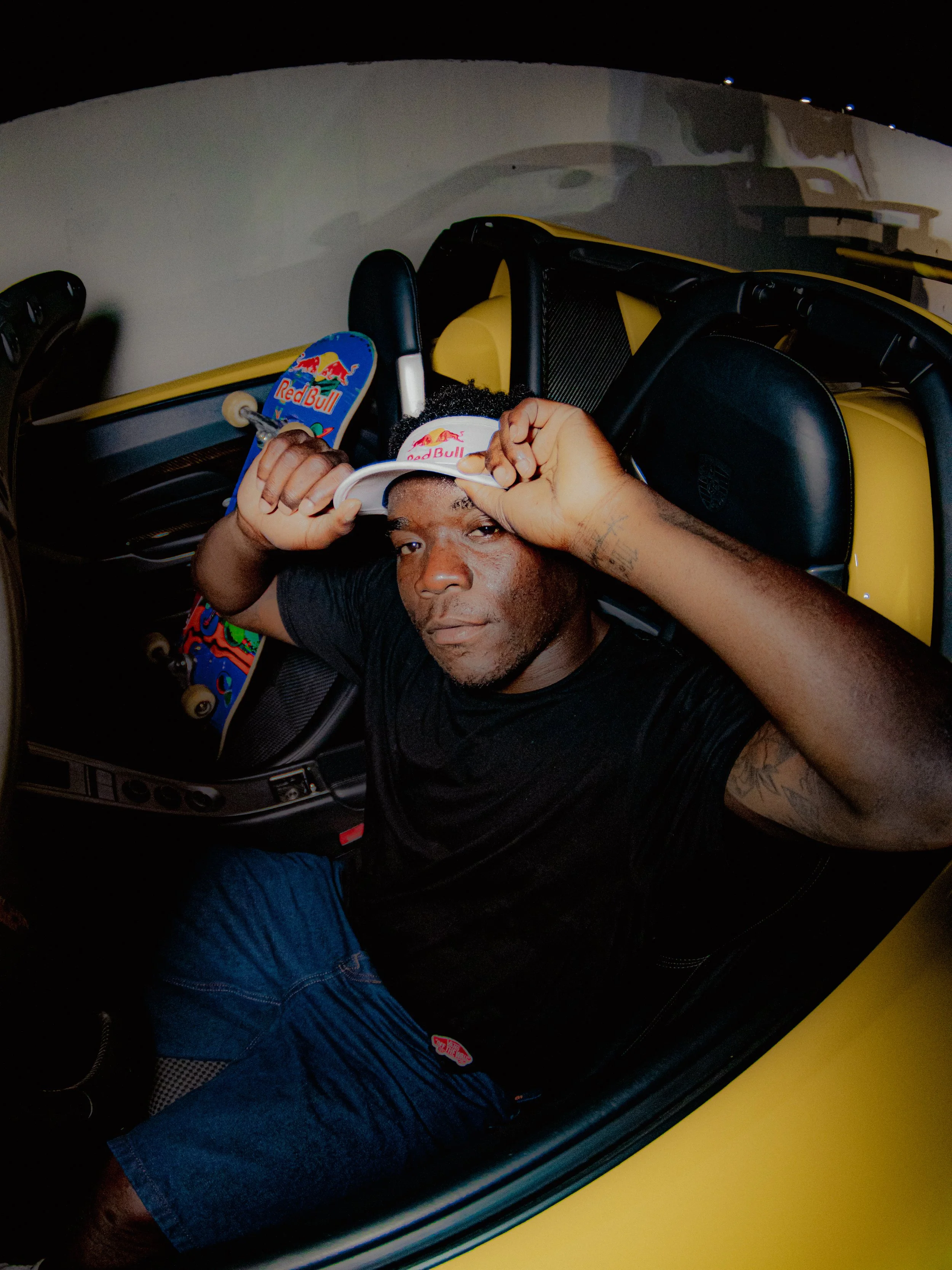 A person sitting in a yellow car with a black interior, holding a Red Bull skateboard above their head, looking at the camera, and wearing a black shirt and blue jeans.