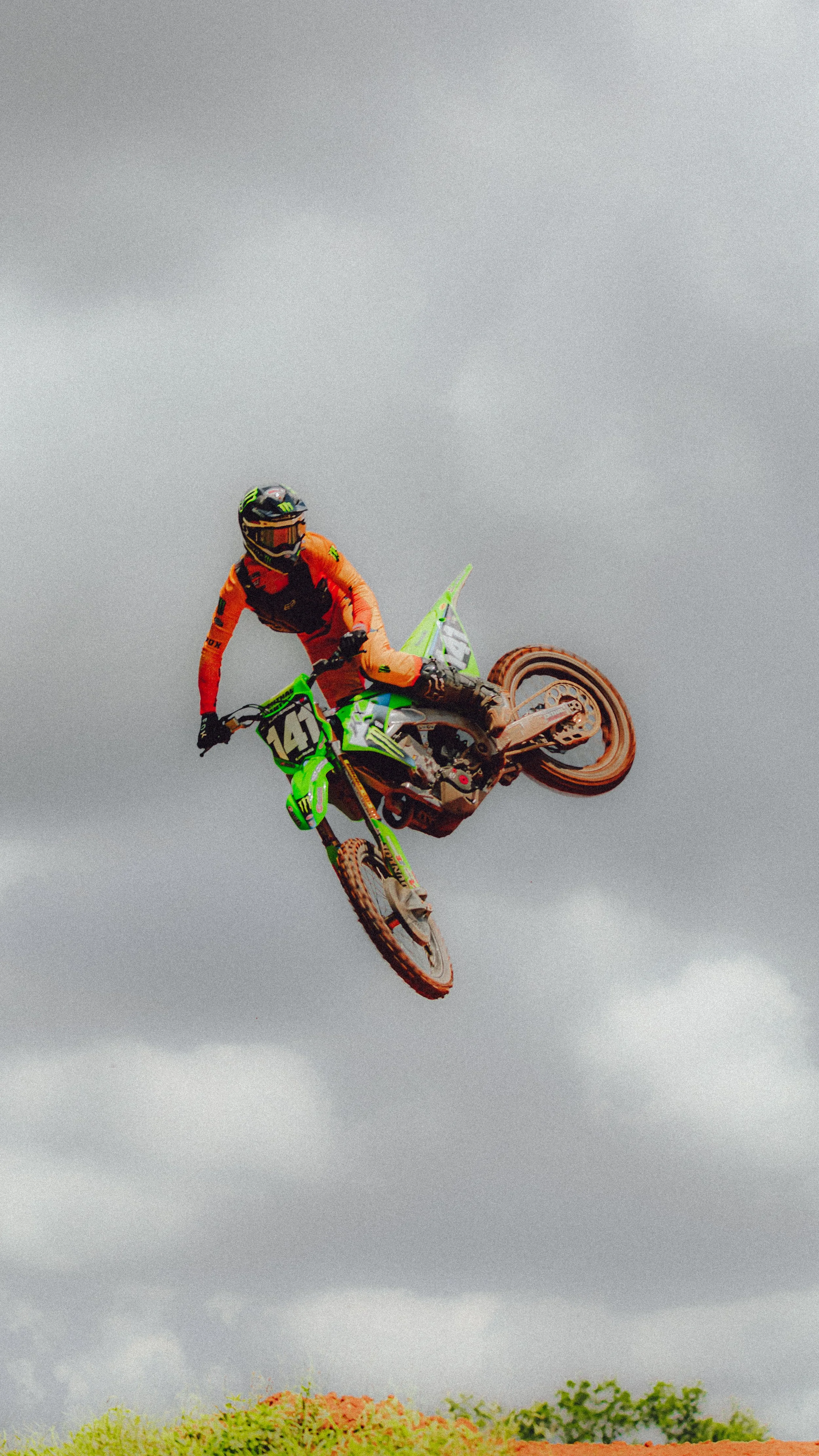 Motocross rider in orange gear and black helmet performing a mid-air stunt on a green and black dirt bike against a cloudy sky.