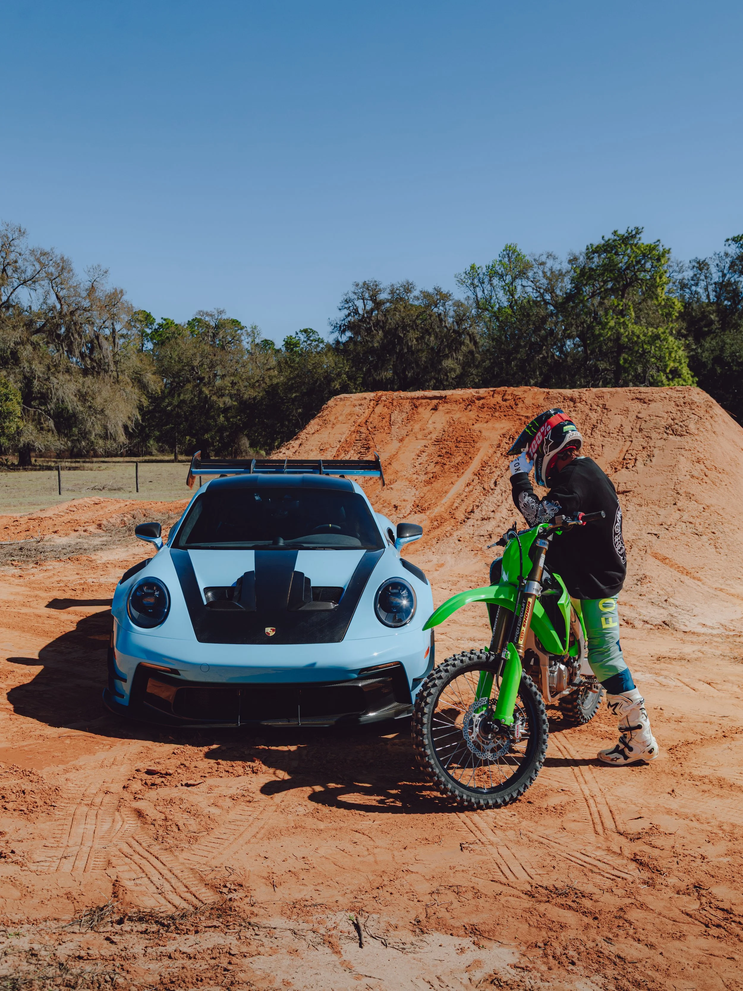 A person in motocross gear standing next to a green dirt bike on sandy terrain, a silver sports car parked nearby, with a large dirt jump and a wooded area in the background under a clear blue sky.