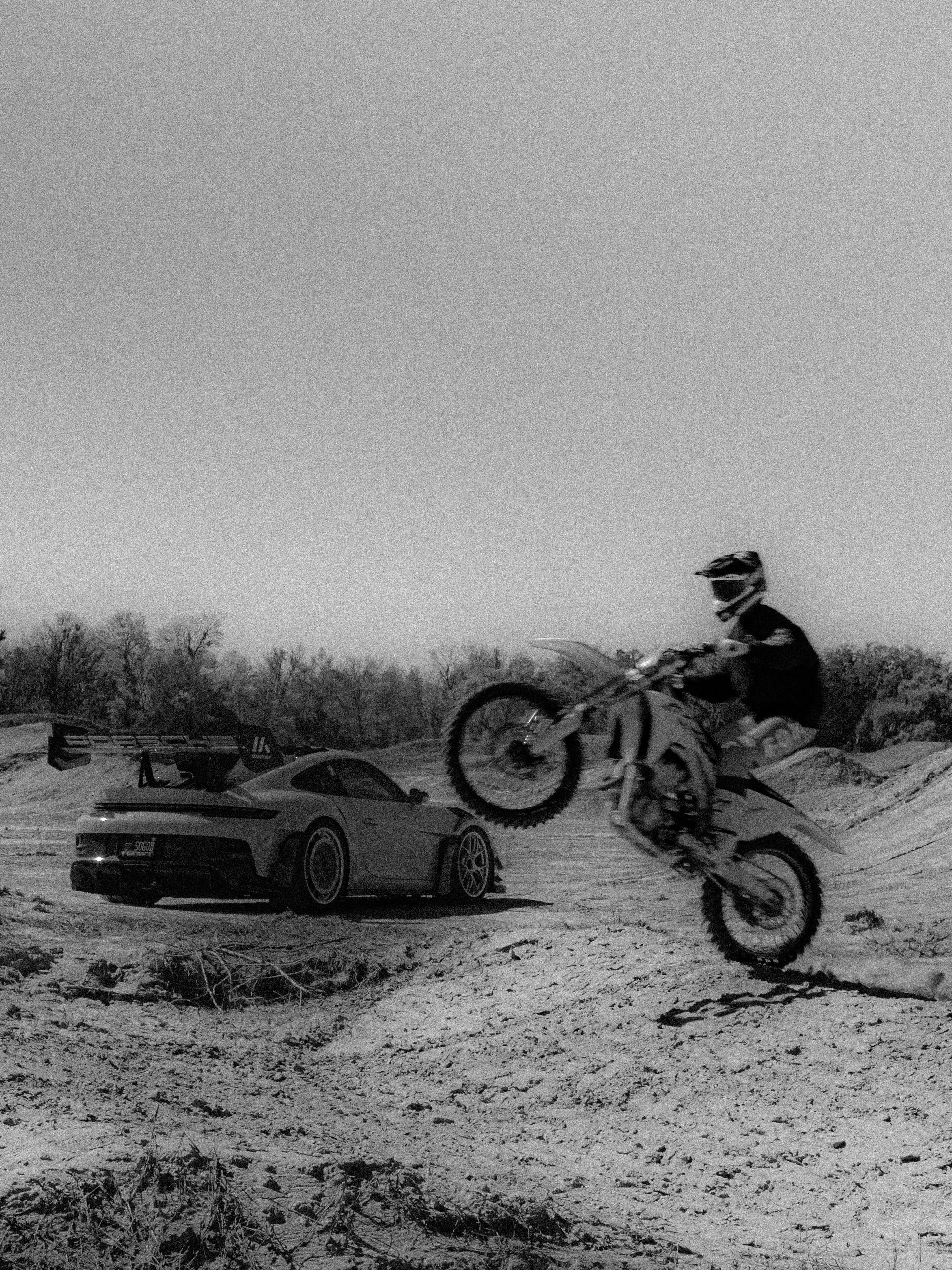 A person riding a dirt bike in a snow-covered outdoor area with a parked car and trees in the background, captured during dusk or nighttime.