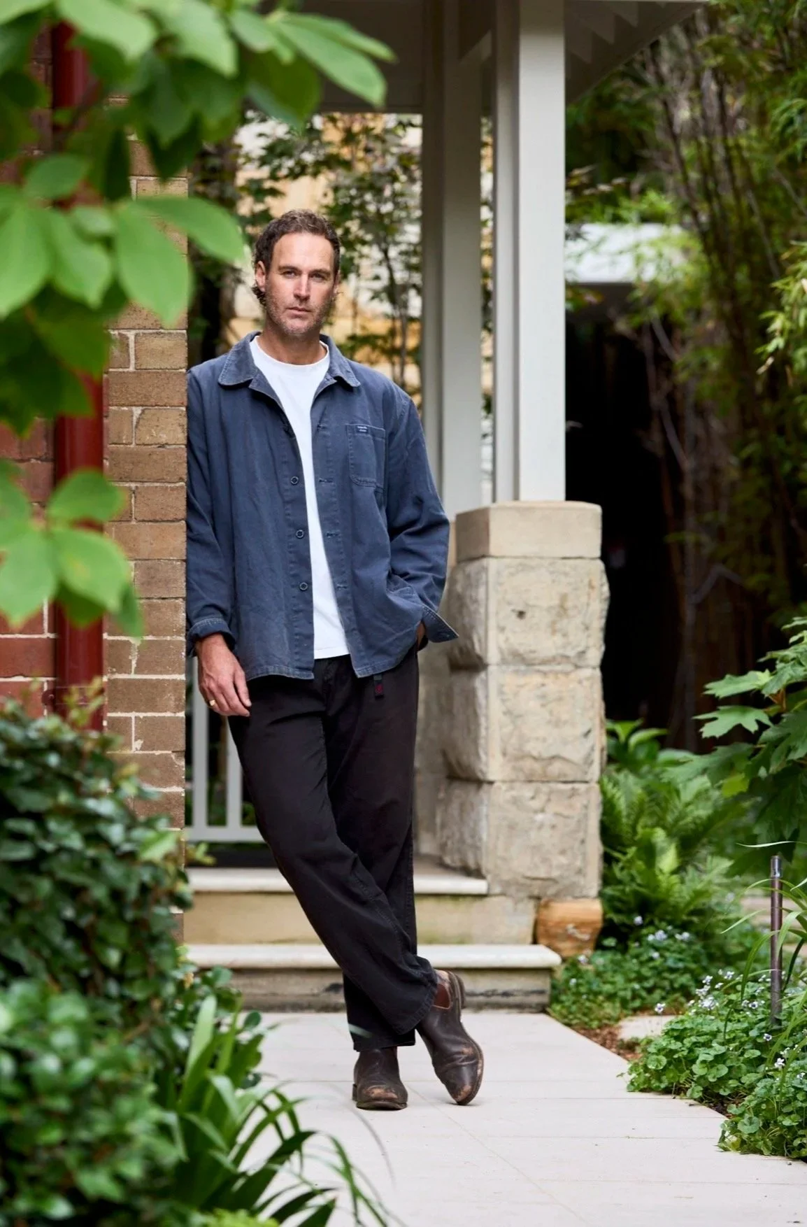 A man leans against a brick wall outdoors with greenery in the background. He is wearing a dark blue button-up shirt over a white t-shirt, black pants, and brown shoes, with his hands in pockets and legs crossed.