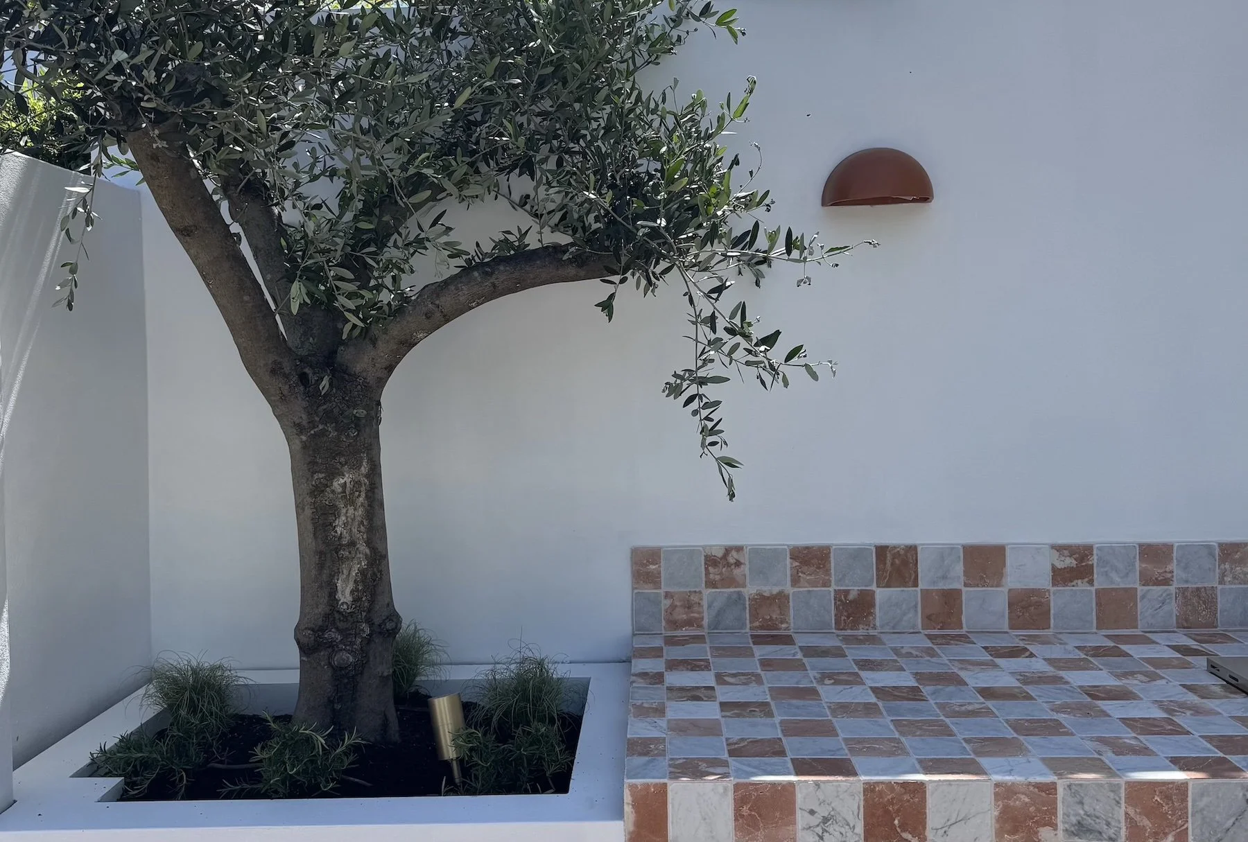 A white wall with a modern half-dome terracotta outdoor wall sconce, a black tree with green leaves, a tiled platform and steps with a mix of red, white, and gray marble tiles, and some small green plants at the base of the tree.