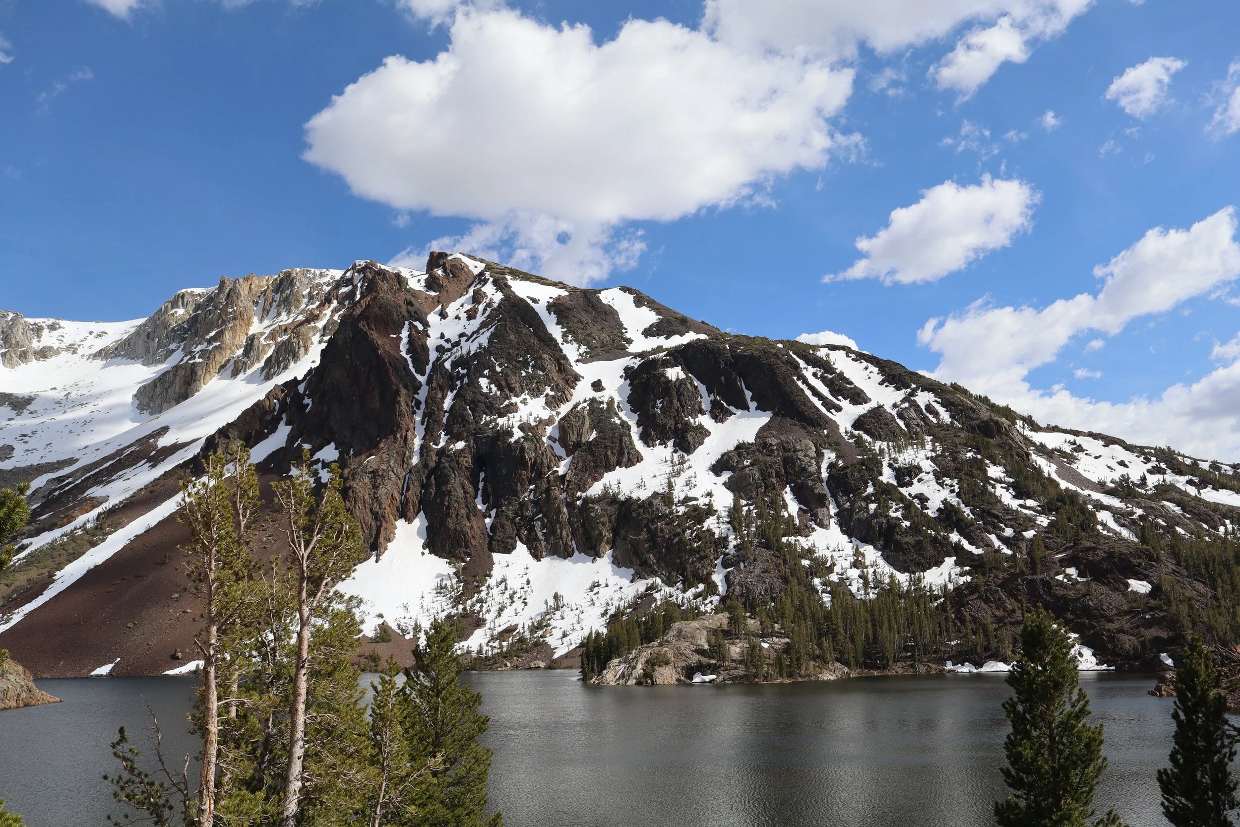 Snow-capped mountain with rocky slopes, trees near a lake, under a partly cloudy sky.
