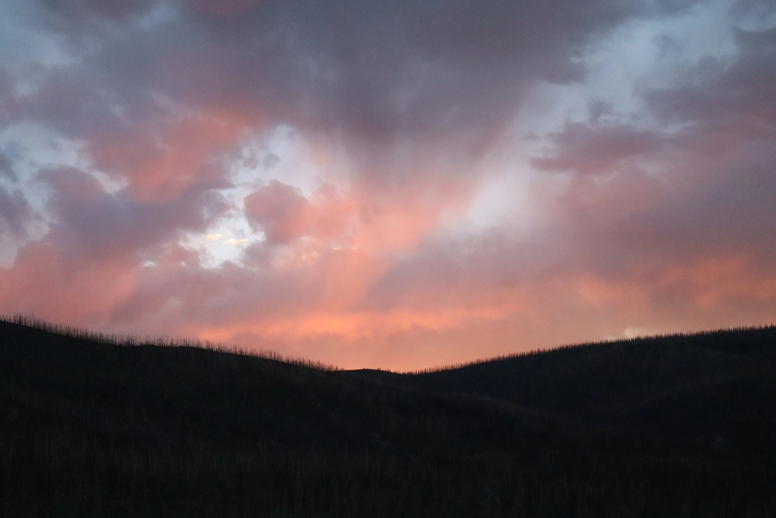 A landscape scene at sunset with a dark silhouette of rolling hills and a colorful sky filled with pink and purple clouds.