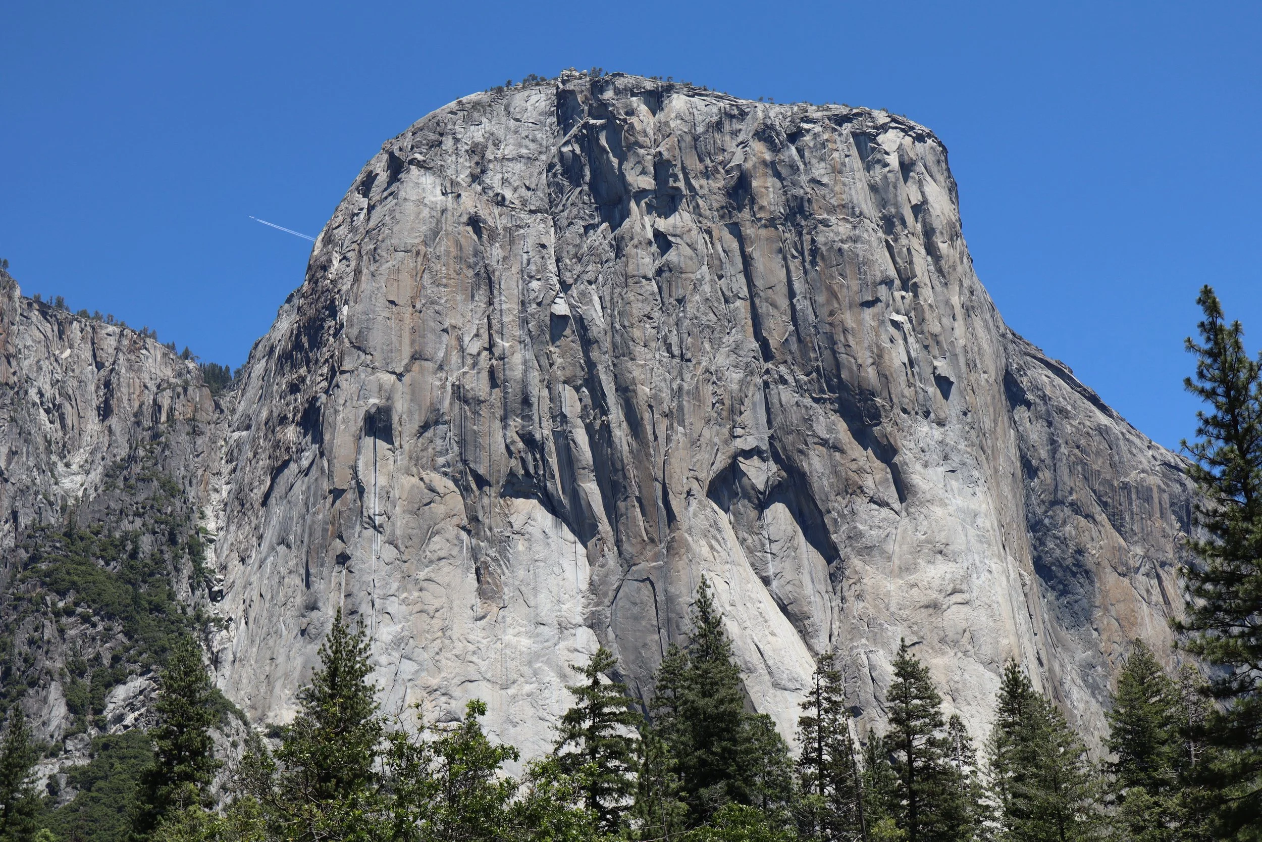 A large granite rock formation with steep, vertical faces and rugged surface, surrounded by green trees and set against a clear blue sky.