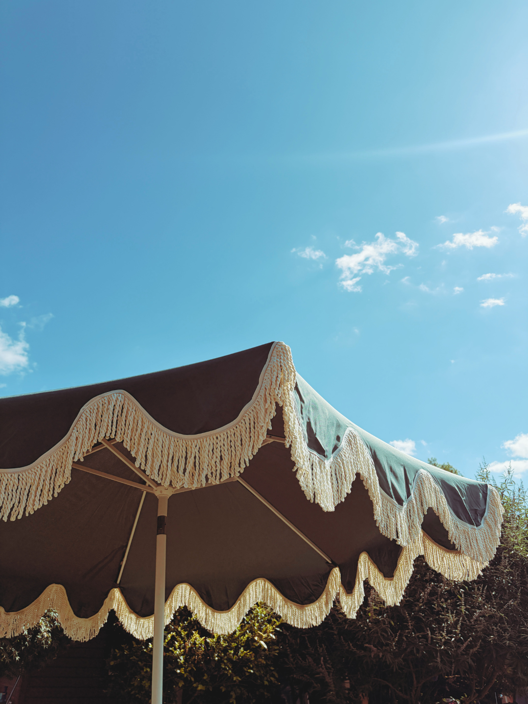An outdoor patio umbrella with scalloped edge and decorative tassels, set against a bright blue sky with a few clouds.