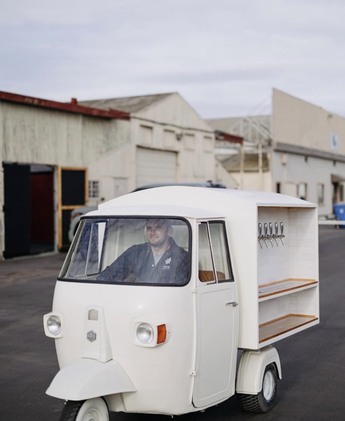 A man driving a small white three-wheeled vehicle, which appears to be a mini food or small merchandise cart, on a street with industrial buildings in the background.