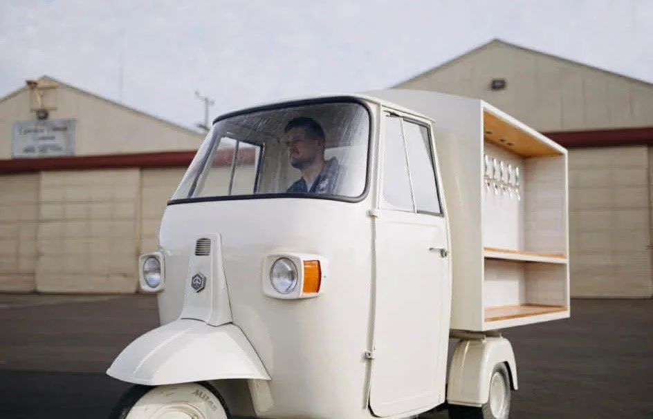 A man driving a small, white, three-wheeled cargo vehicle with a large wooden storage compartment in the back, in an outdoor setting.