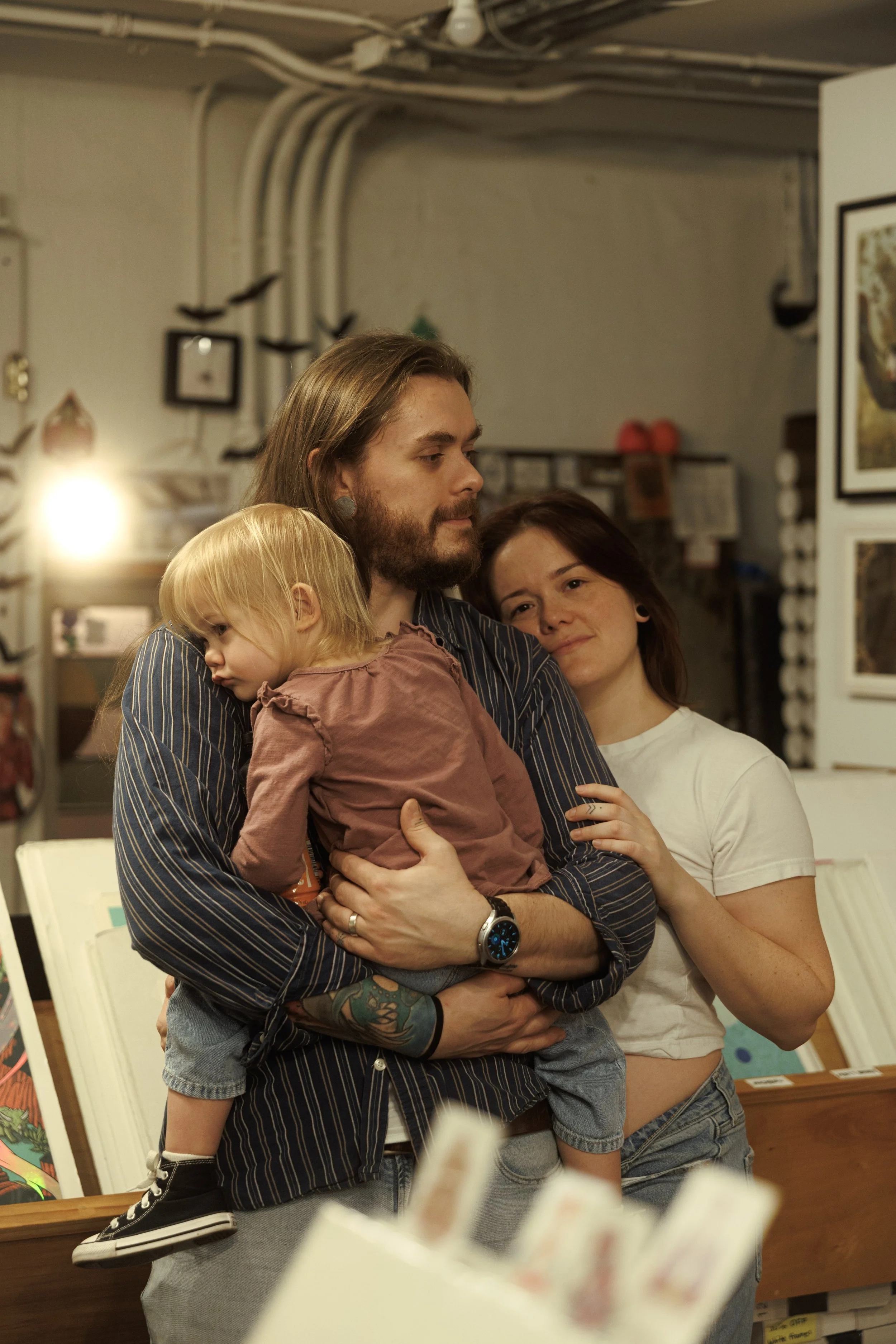 A group of three young people, a man, a woman, and a child, are embracing and posing together inside a store or gallery with artwork on the walls. The man has long hair, a beard, and tattoos on his arm, and is wearing a striped shirt. The woman has shoulder-length hair and is wearing a white t-shirt. The child has blonde hair and is wearing a striped shirt with sneakers.