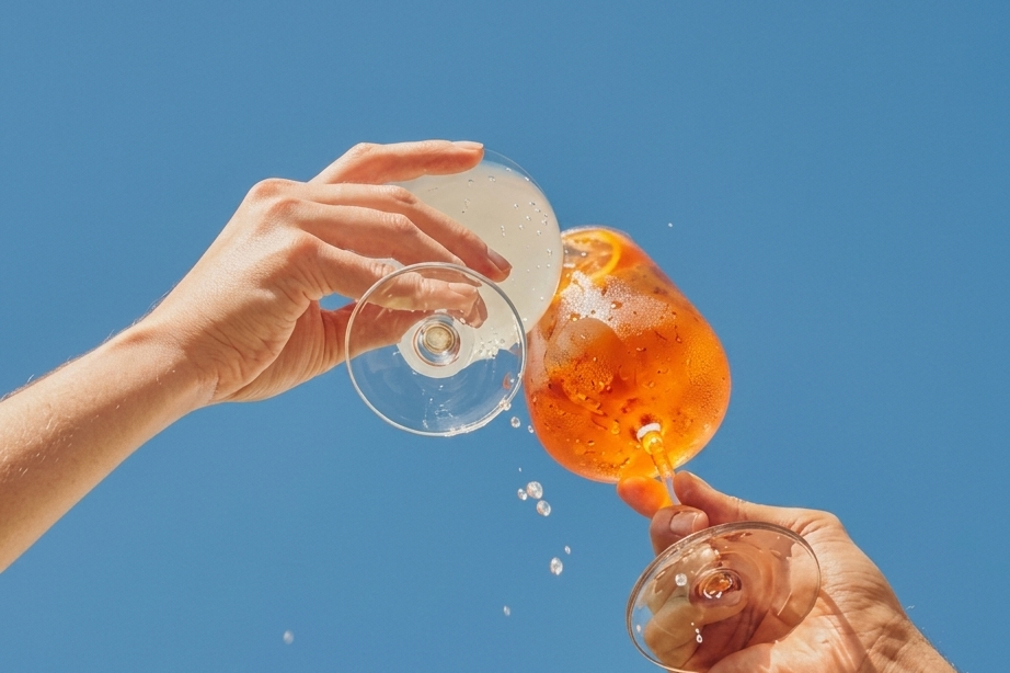 Two people clinking glasses filled with cocktails against a blue sky, one with an orange slice and the other with a lemon slice.
