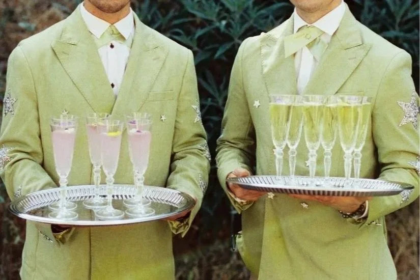 Two people holding silver trays with glasses of champagne and a pink cocktail, dressed in matching light green suits with star embellishments.