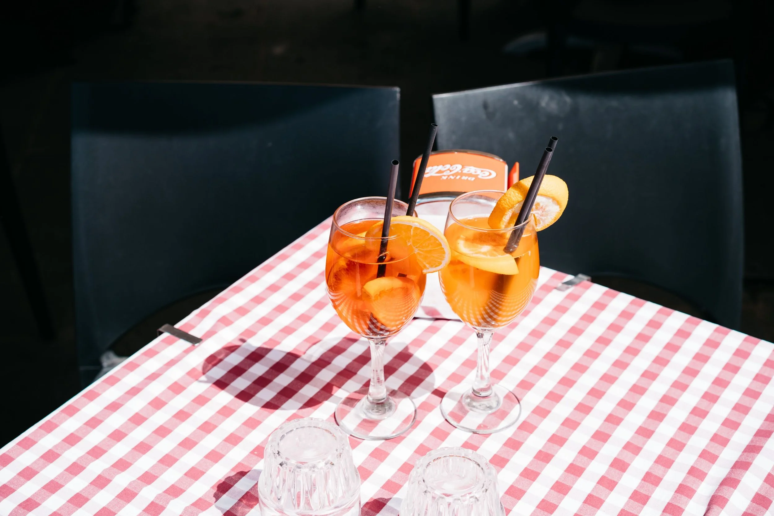 Two glasses of orange-colored drinks garnished with lemon slices and black straws, placed on a red and white checkered tablecloth.