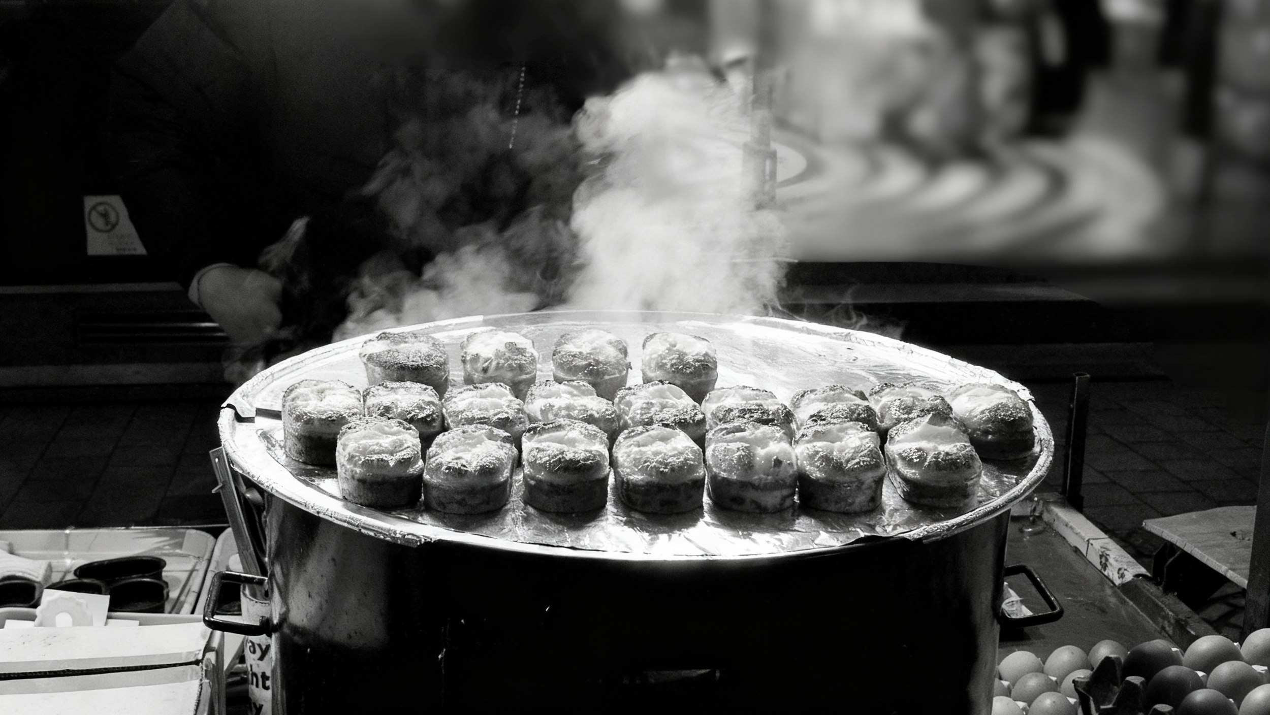 A street vendor is cooking round pastries with powdered sugar on a large tray, with steam rising from the hot pastries.