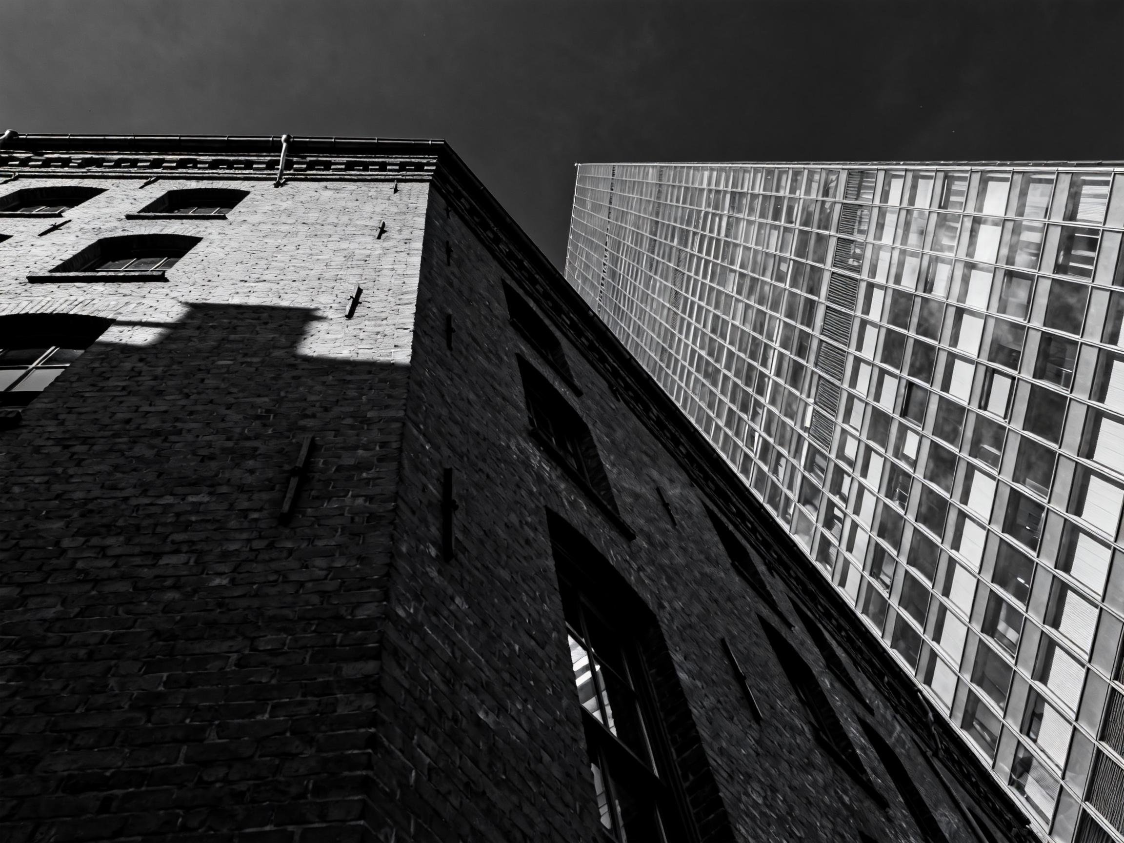 Low-angle view of contrasting architecture with an old, textured brick building on the left and a modern glass skyscraper on the right, against a dark sky.