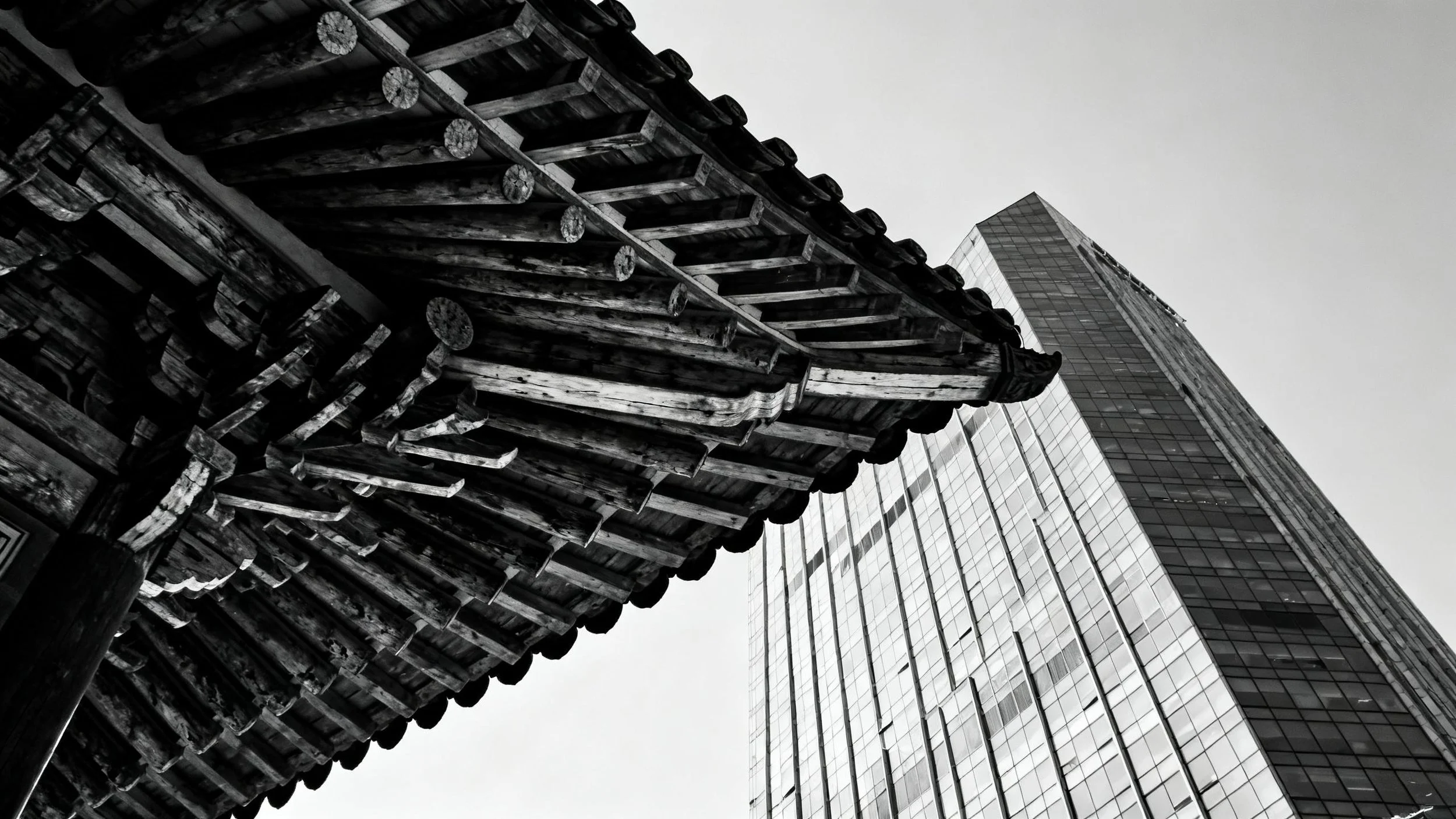A low-angle black-and-white photograph of two contrasting buildings: a traditional wooden pagoda-style structure in the foreground and a modern glass skyscraper in the background.