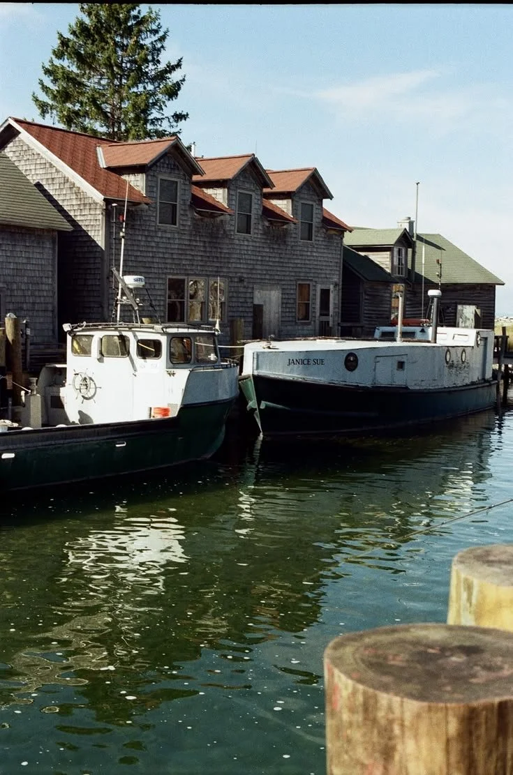 Two boats docked along a wooden pier in front of weathered seaside houses, with calm water reflecting the structures and boats.