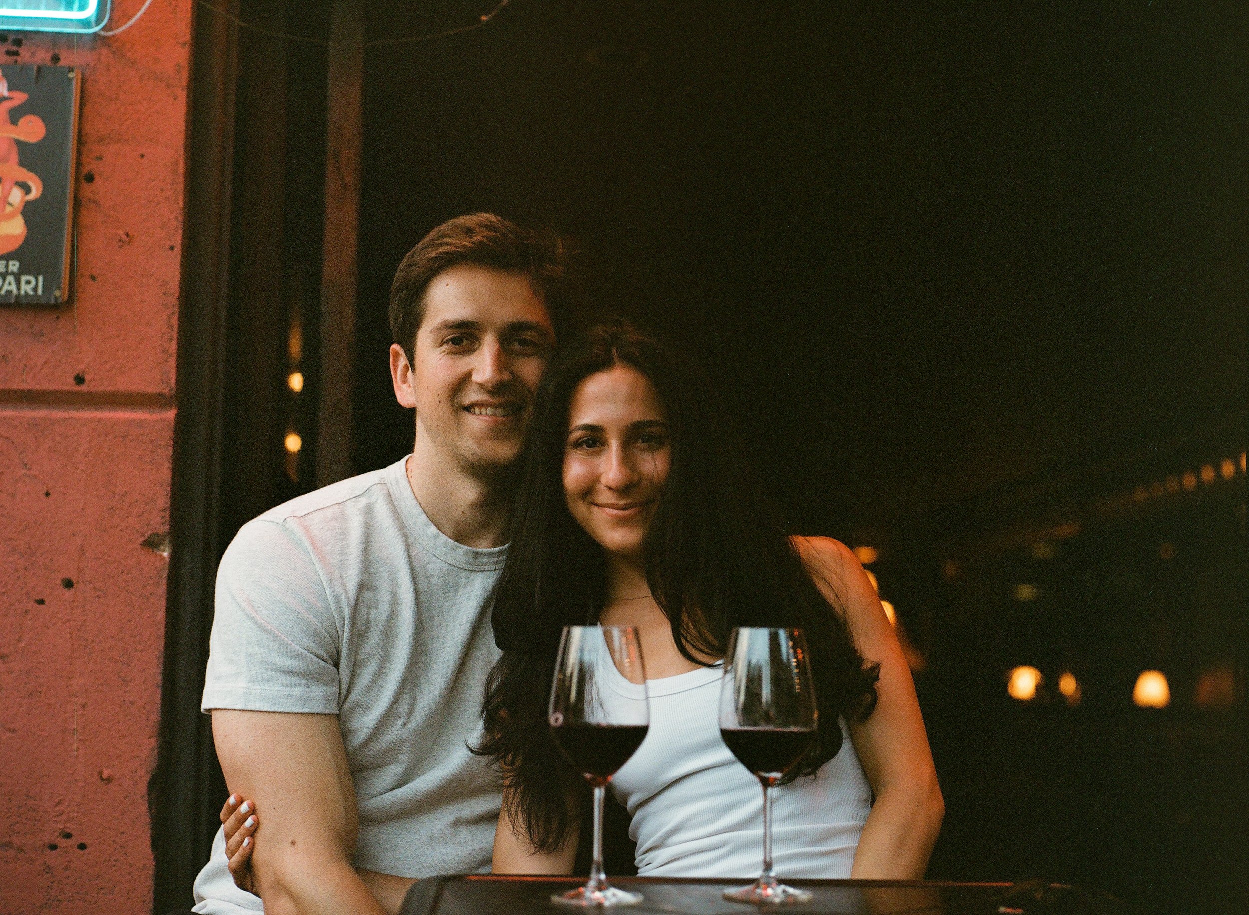 A young man and woman sitting at a restaurant table with glasses of red wine in front of them, smiling and leaning close together.