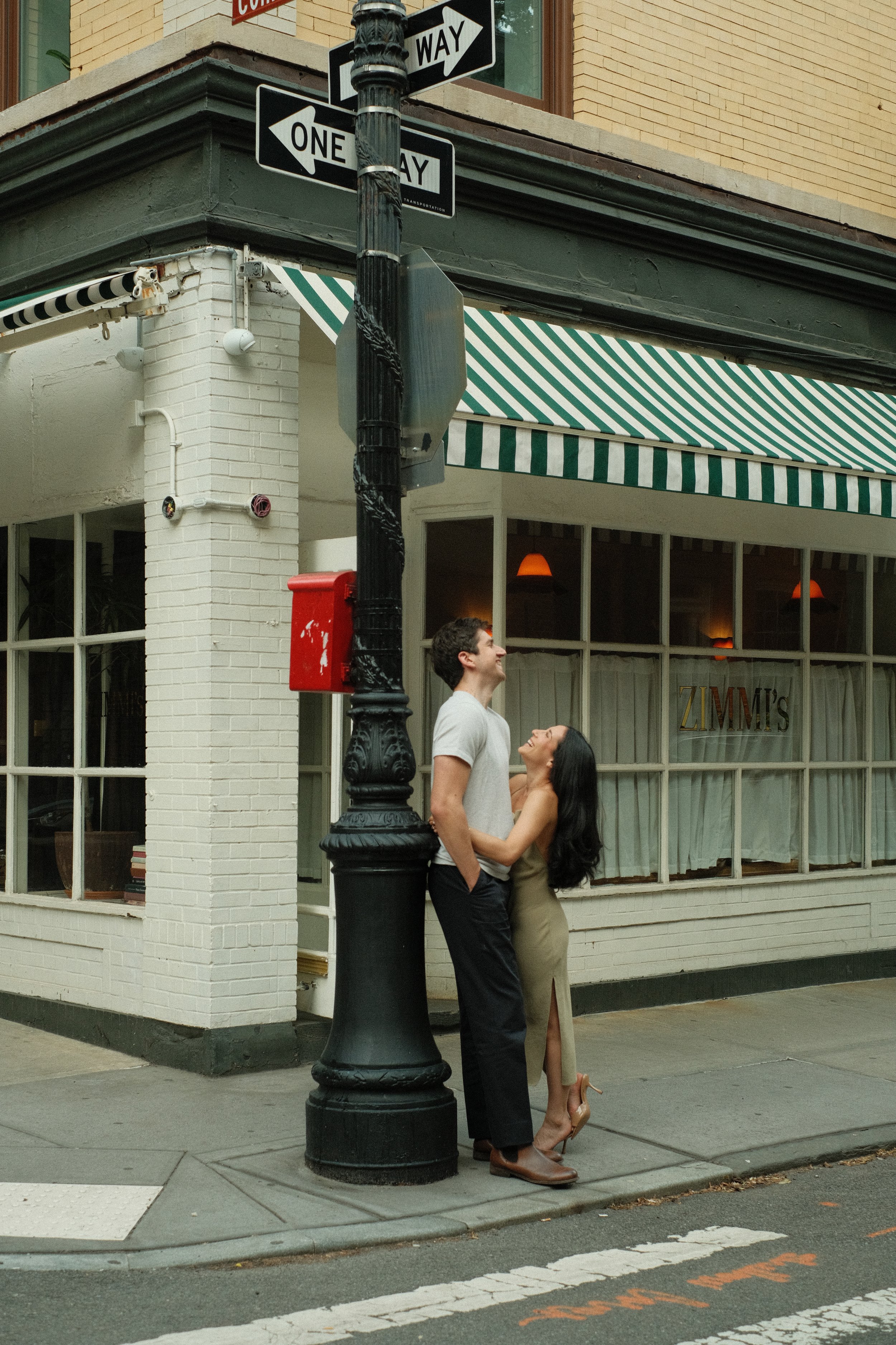 A couple standing on a city sidewalk, embracing and looking at each other, with a black lamppost and a corner of a building with green and white striped awning and large windows behind them.