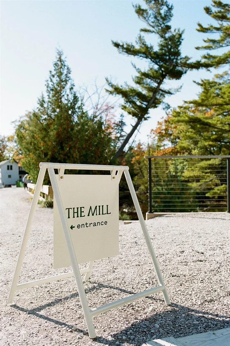 A white A-frame sign with green text reading 'The Mill entrance' and an arrow pointing left, set on gravel with trees and a fence in the background.