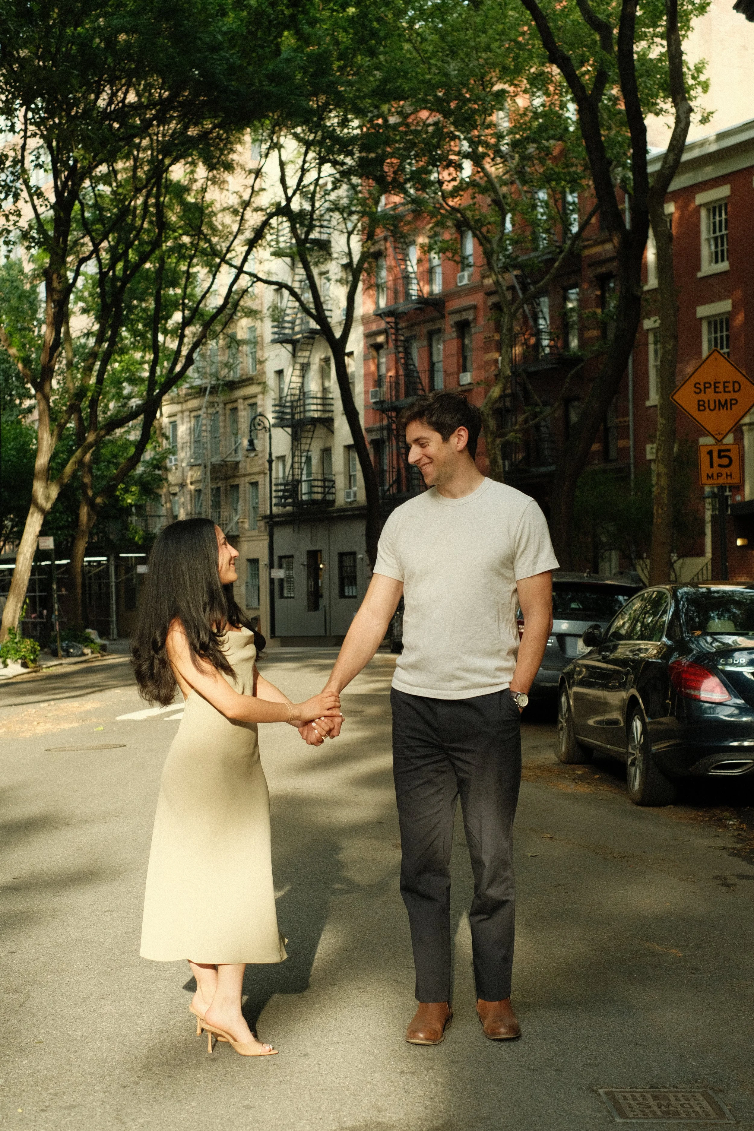 A man and a woman holding hands and smiling at each other on a city street. The woman is wearing a beige dress and high heels, while the man is wearing a light shirt, dark pants, and brown shoes. Trees and apartment buildings are visible in the background, along with parked cars and a yellow traffic sign indicating a speed bump with a 15 miles per hour speed limit.