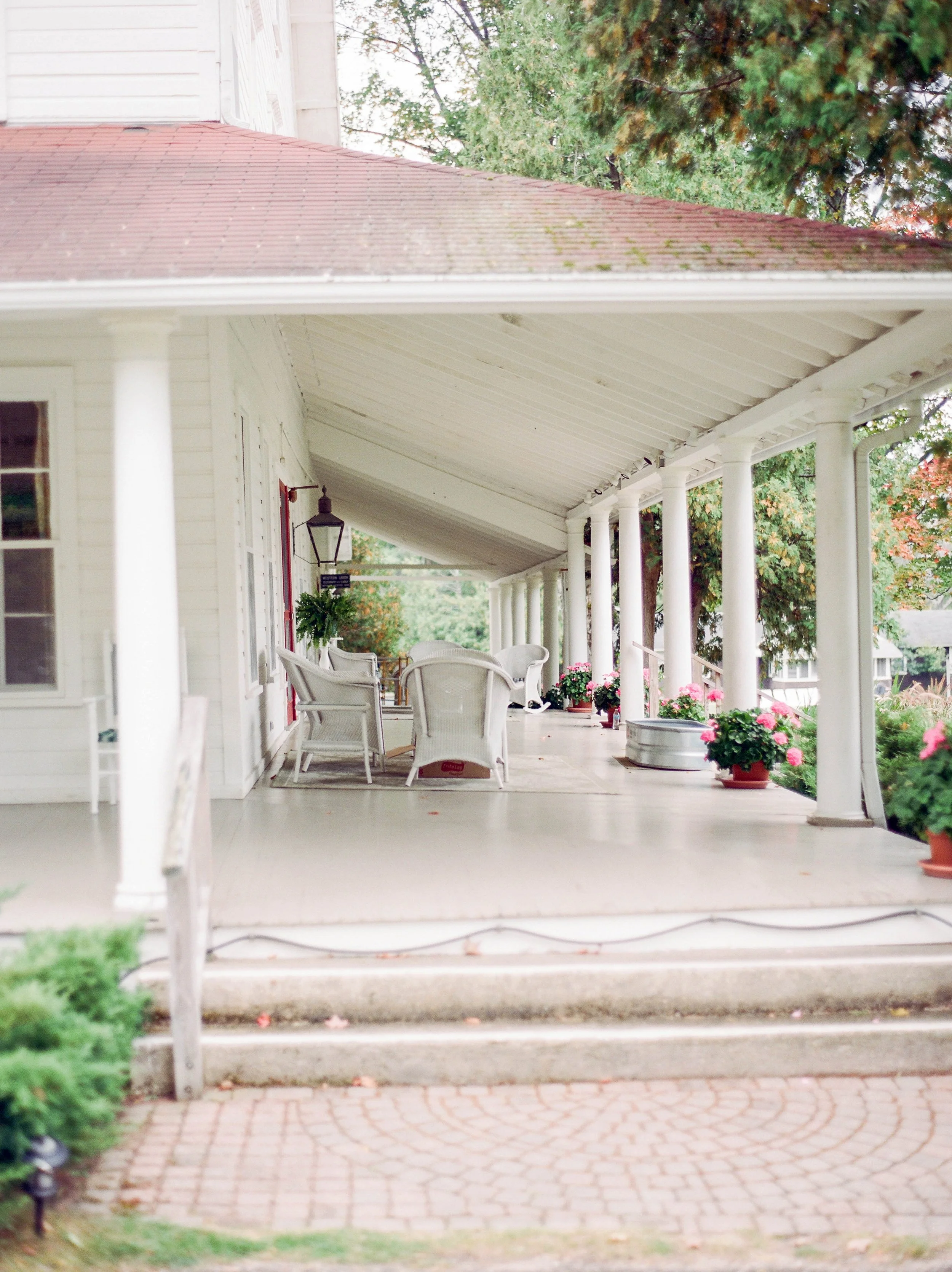 Porch with white chairs, potted flowers, and a hanging plant, supported by white columns, with trees in the background.