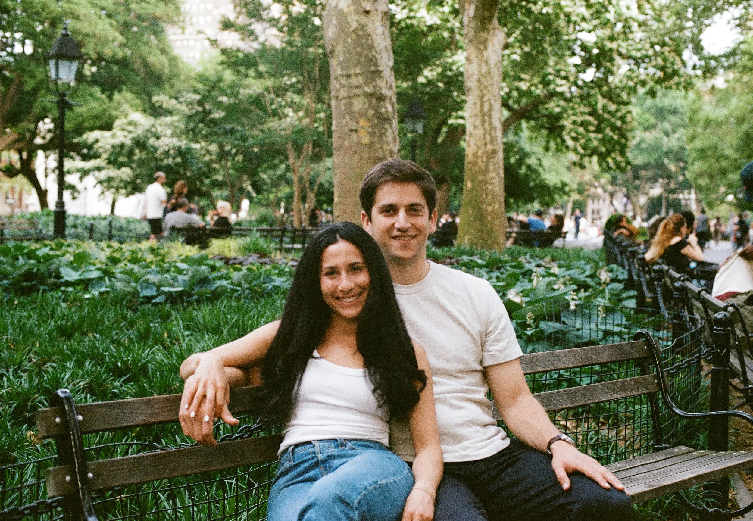 A young woman with long dark hair and a young man with short dark hair sitting closely on a park bench, smiling at the camera. They are surrounded by greenery in a park, with other people sitting on benches and walking in the background.
