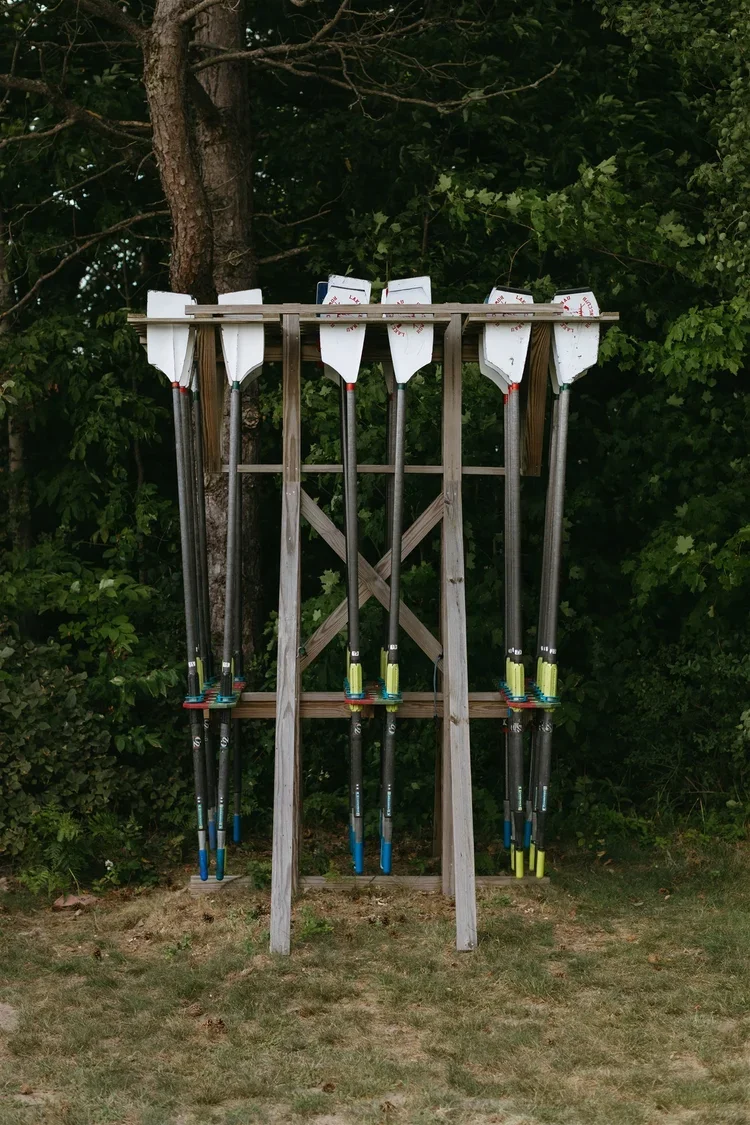 Several snow shovels hanging on a wooden rack outdoors, with green trees in the background.