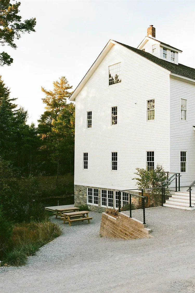 A white multi-story house with a stone foundation, black railings, and multiple windows, surrounded by trees with a gravel pathway and picnic table outside.