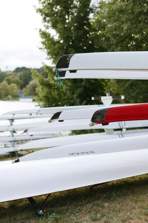 Multiple canoes or kayak boats stacked on racks outdoors with trees and water in the background.