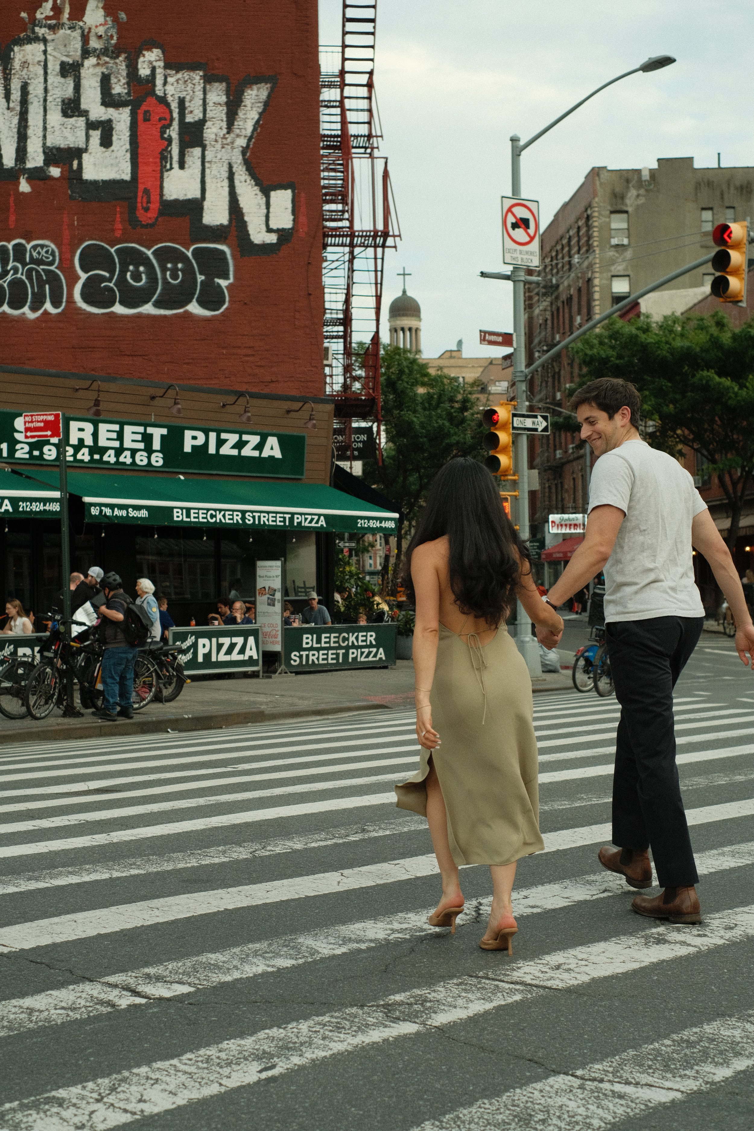 A man and a woman holding hands and crossing a street at a crosswalk in a city. The woman is wearing a beige dress and high heels, while the man is in black pants and a white shirt. There are buildings, a pizza restaurant sign, and traffic lights in the background.