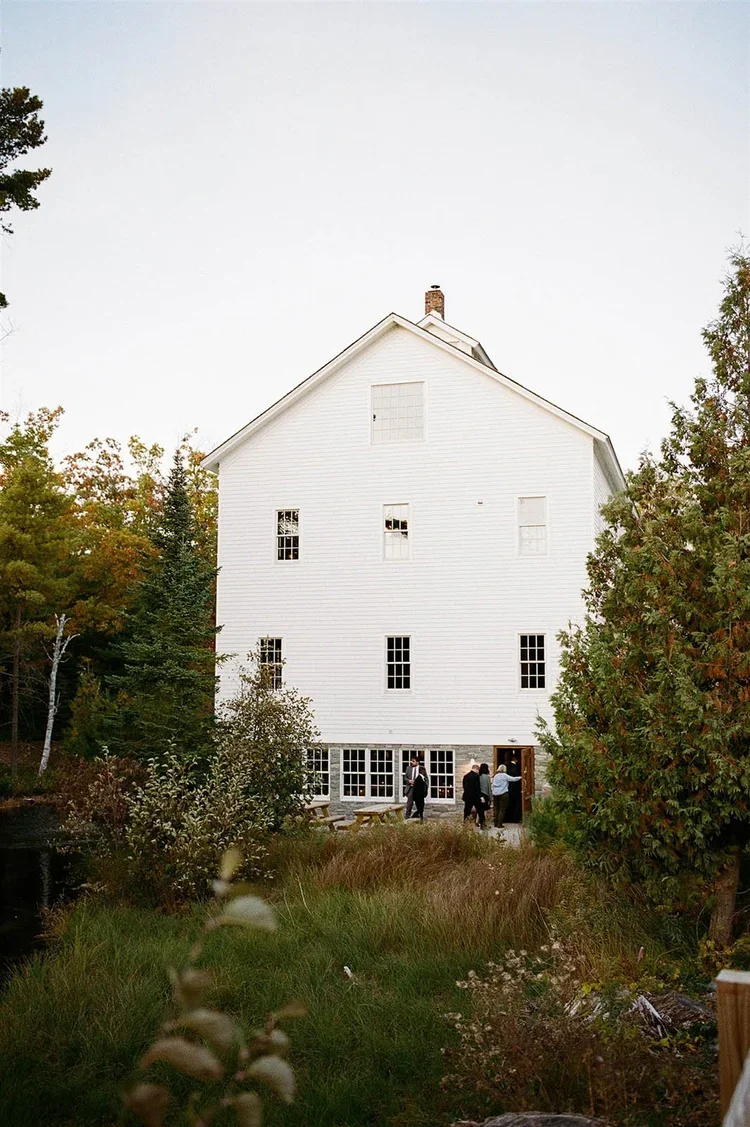 A white multi-story building with several windows, surrounded by trees and greenery, with a group of people gathered near the entrance.