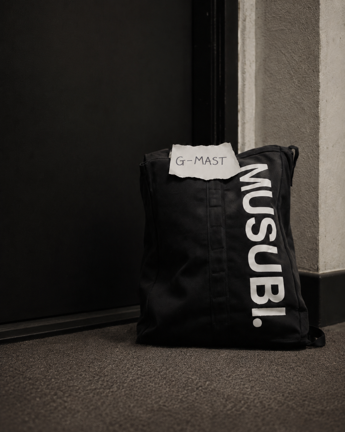 A black duffel bag with white text on the side and a torn piece of paper with "G-MAST" written on it, placed on the floor near a wall and door.