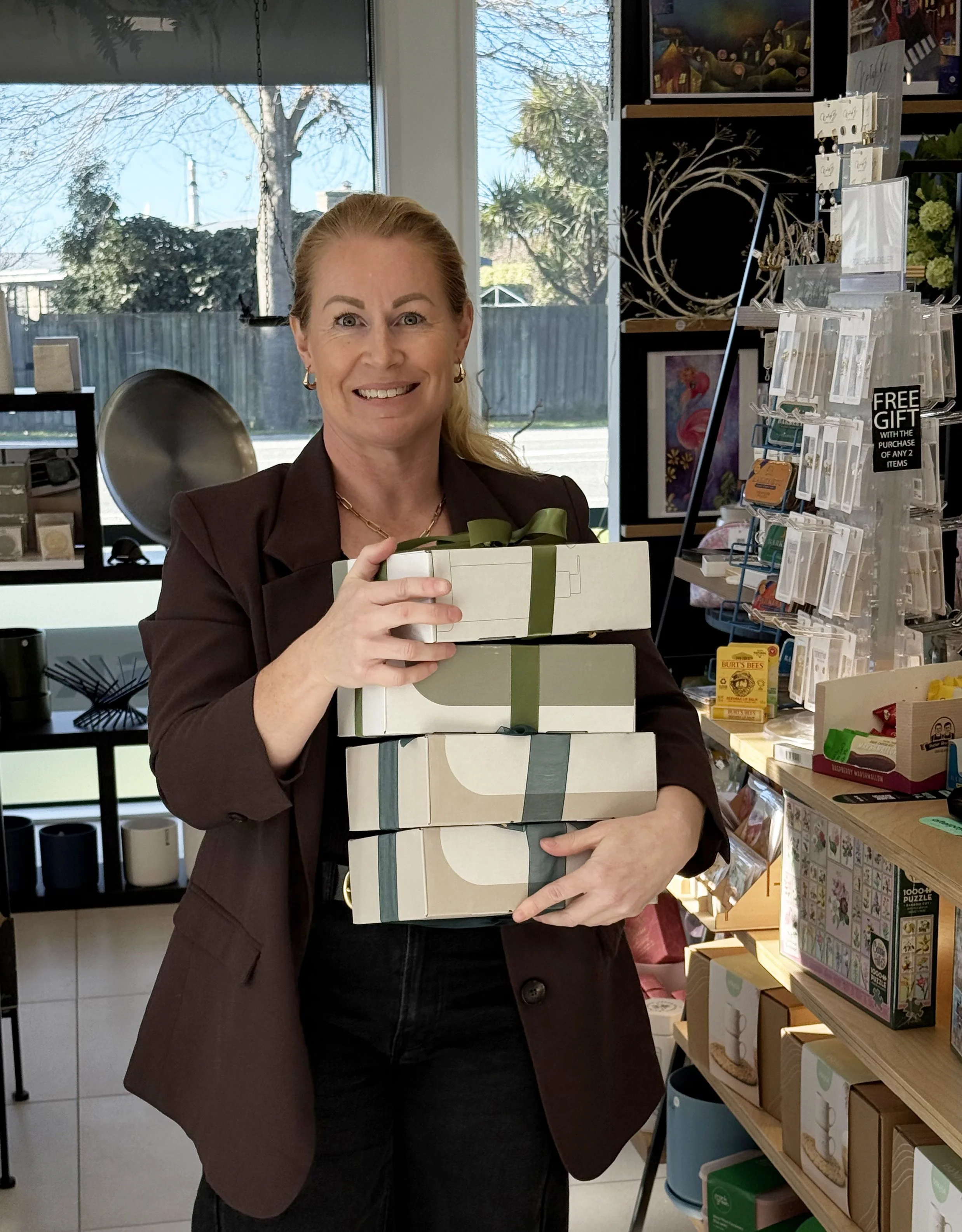 Hannah Brown holding a stack of gift boxes decorated with ribbons inside a store with shelves of various products.