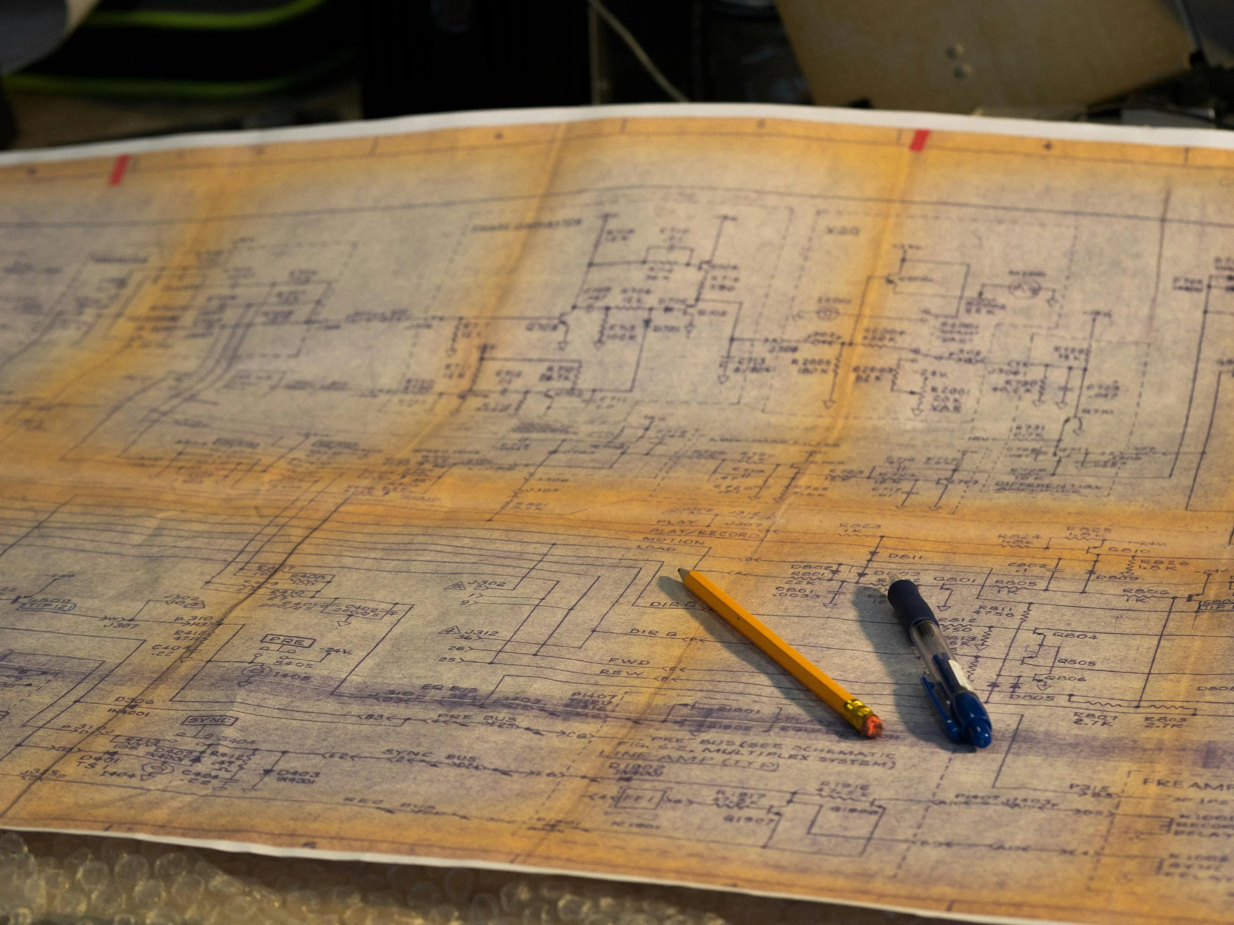 A detailed engineering blueprint laying on a table with a yellow pencil, a black pen, and a blue pen placed on top of it.