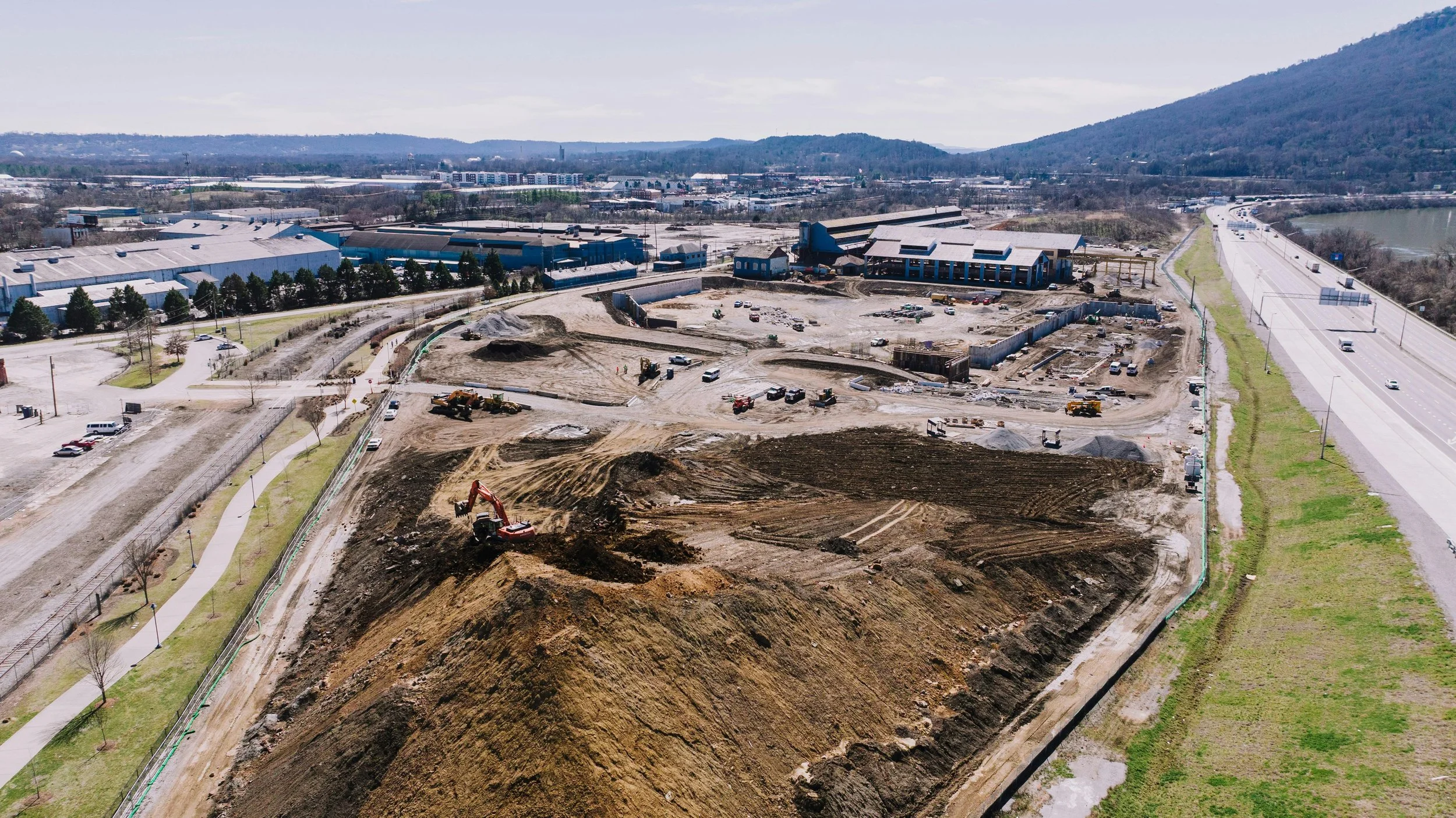 Construction site with heavy machinery and dirt piles, near a road and industrial buildings, with mountains in the background.